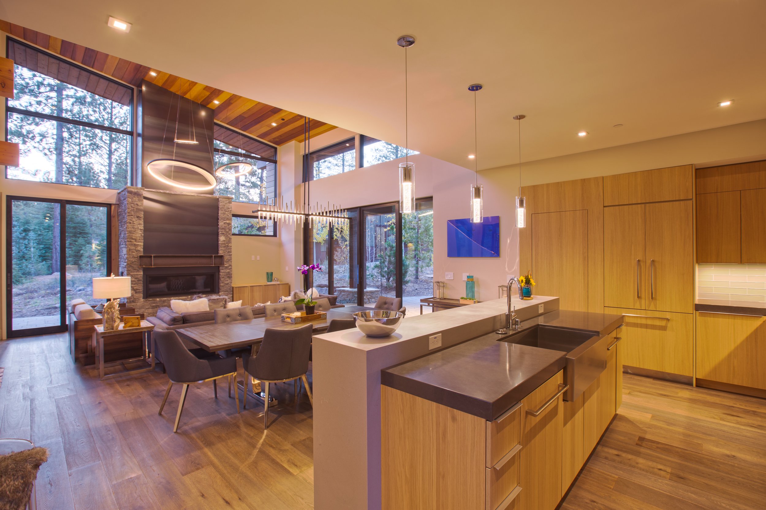 Open-concept living and kitchen area with wooden cabinets, large windows, dining table, modern lighting, and a fireplace.