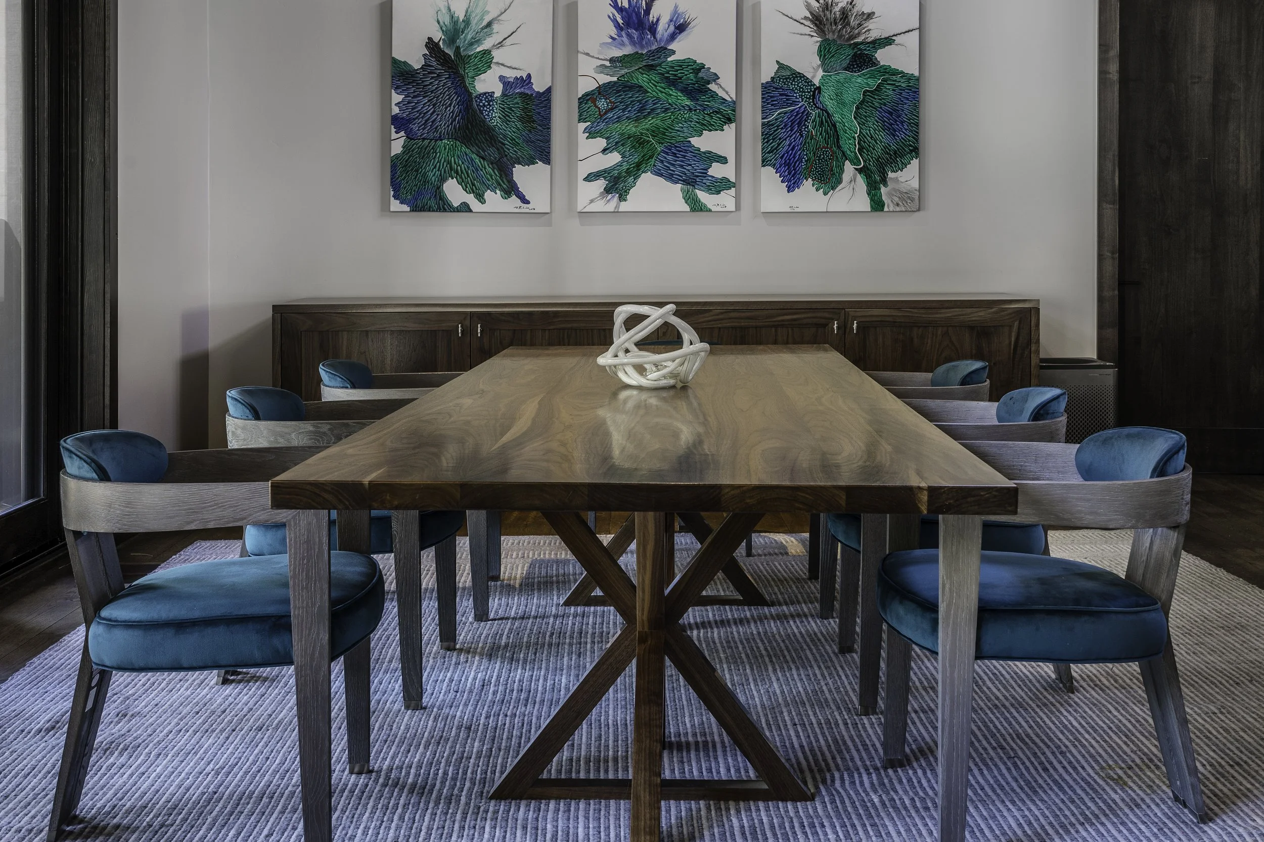 A rectangular wooden dining table with a decorative white sculpture in the center, surrounded by nine chairs with blue upholstered seats, in front of three abstract botanical paintings with blue and green hues on a white wall, and a rug underneath.