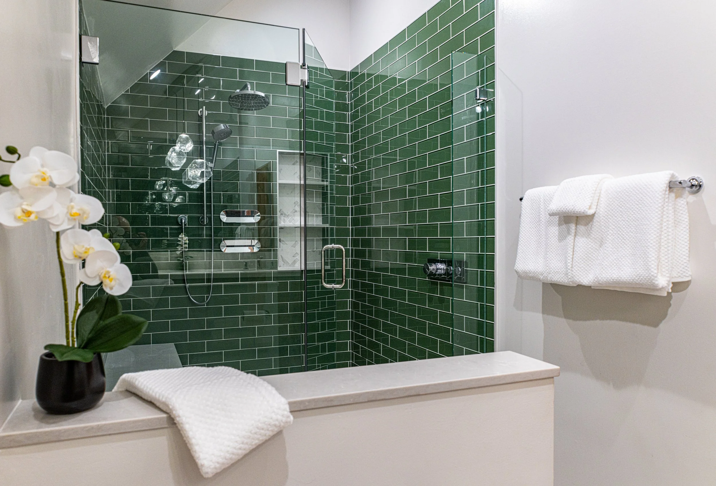 A modern bathroom featuring a walk-in shower with green subway tile walls, a glass door, and multiple showerheads. There is a white towel on a bar, a towel on a ledge, and a potted orchid on the ledge.