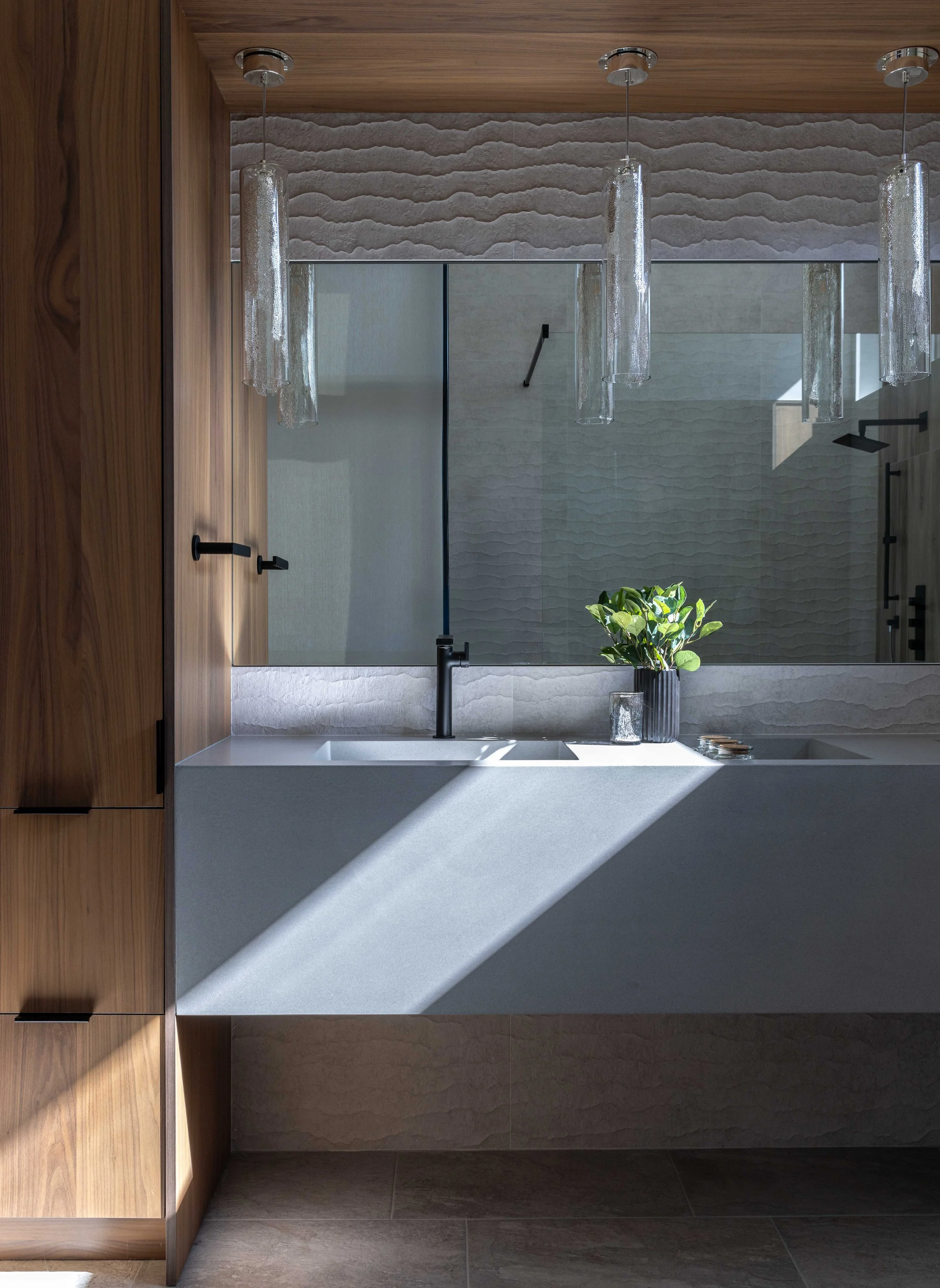 Modern bathroom with a floating white sink, black faucet, large mirror, wooden cabinets, hanging glass pendant lights, green plant in a black vase, and a shower with a black showerhead in the background.