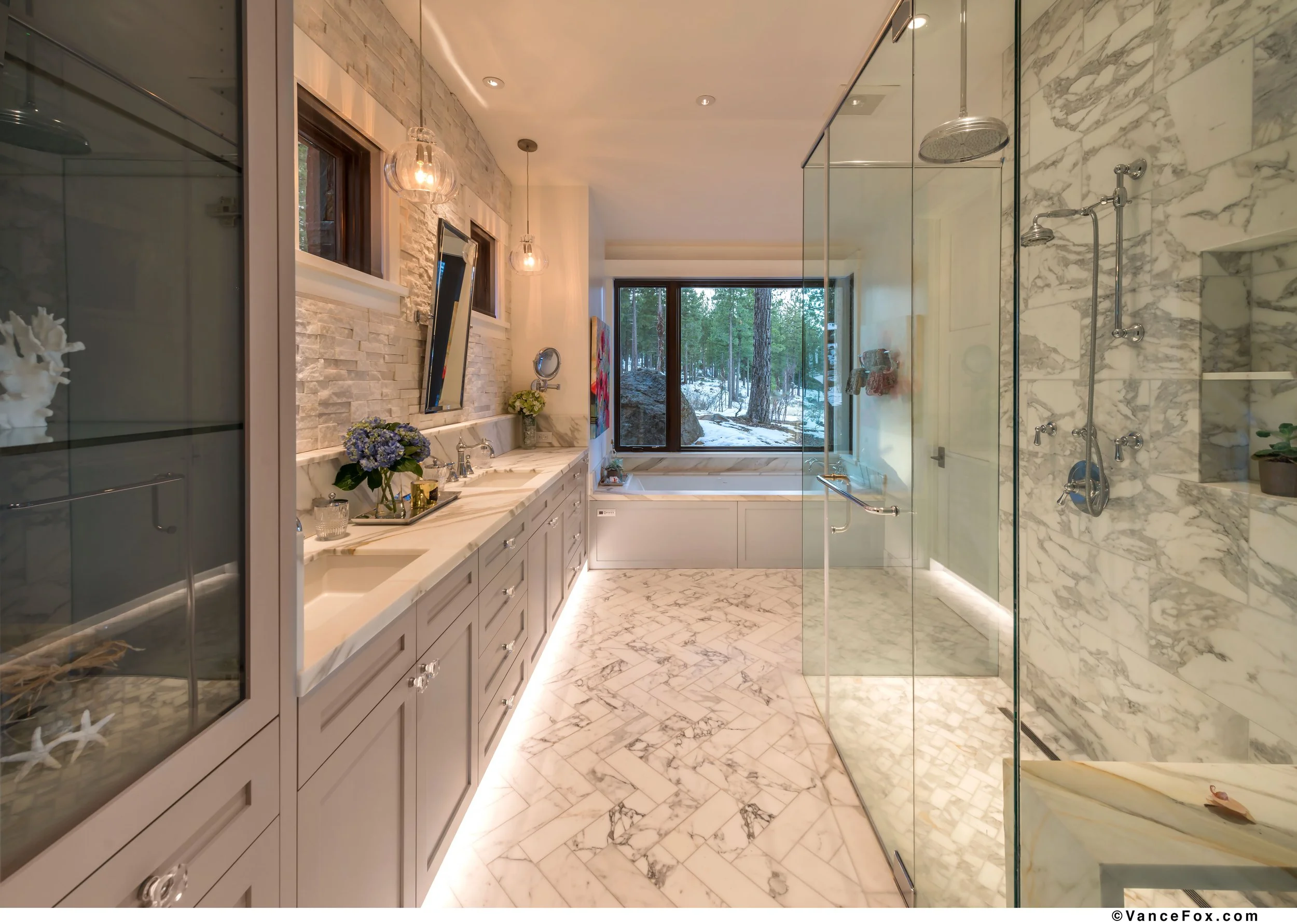 Luxurious bathroom with marble floors and walls, a large bathtub by a window with forest view, double sink vanity with flowers and decorative items, glass-enclosed shower area, and ambient lighting.