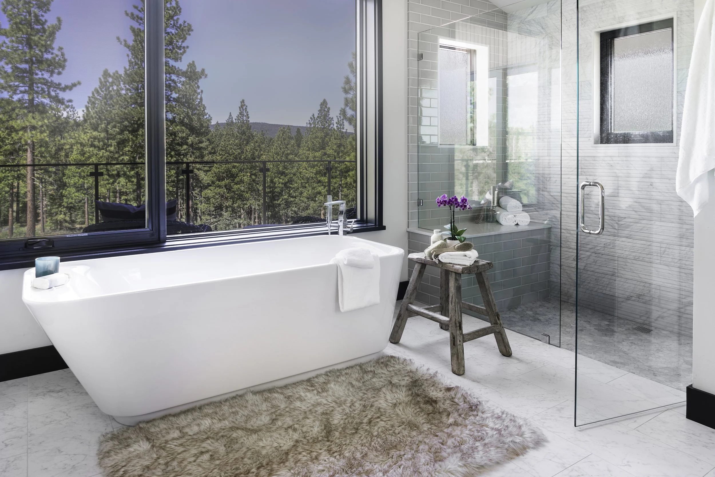 Modern bathroom with a large bathtub, a glass-enclosed shower, and a large window overlooking a forested landscape. The bathtub has a towel draped over the edge, and there's a small wooden stool holding rolled towels and a potted purple orchid. The b