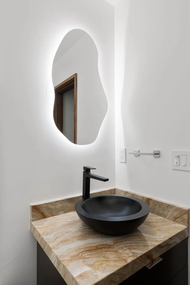 Modern bathroom sink with a black vessel basin, black faucet, marble countertop, round mirror with backlit lighting, and white walls.