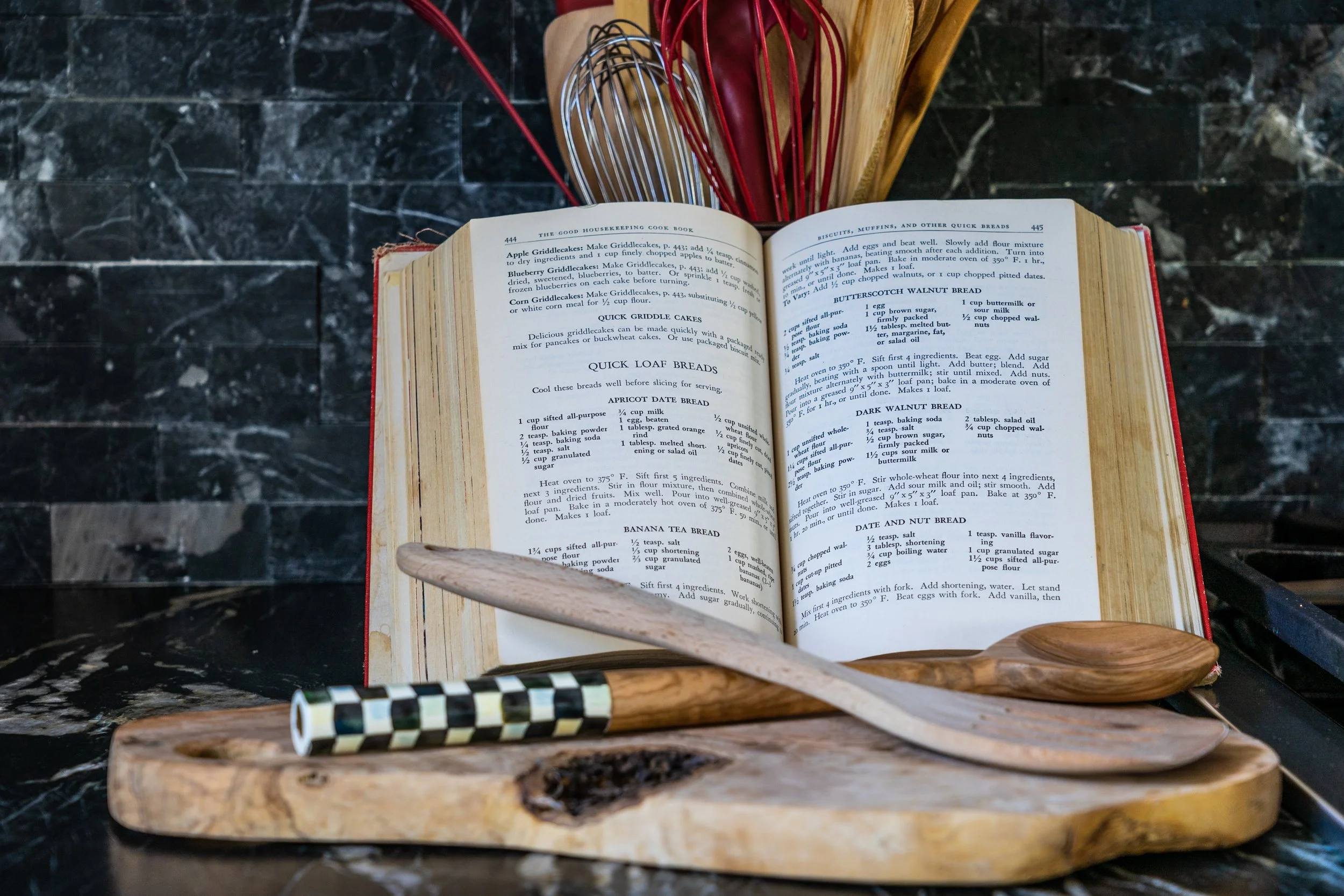 Open cookbook with recipes for bread, placed on a kitchen countertop, surrounded by various wooden kitchen utensils and utensils holders.