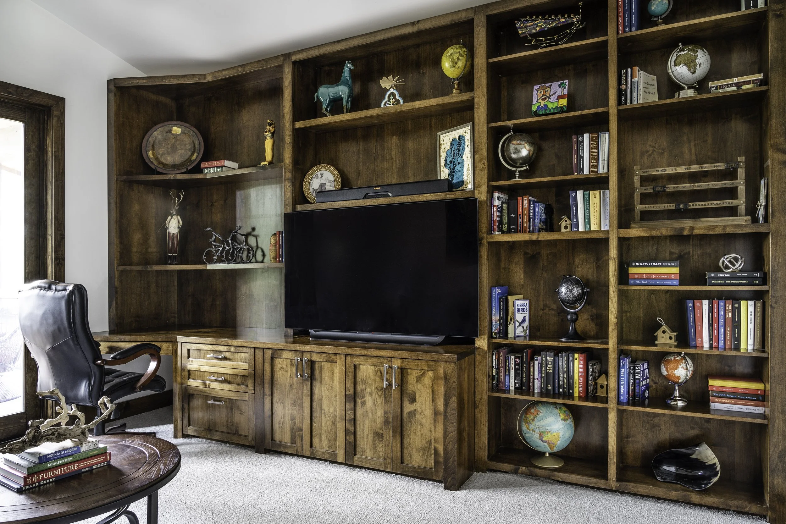 Wooden built-in bookshelf with decorative globes, books, and art pieces, with a large flat-screen TV in the center and a black office chair nearby.