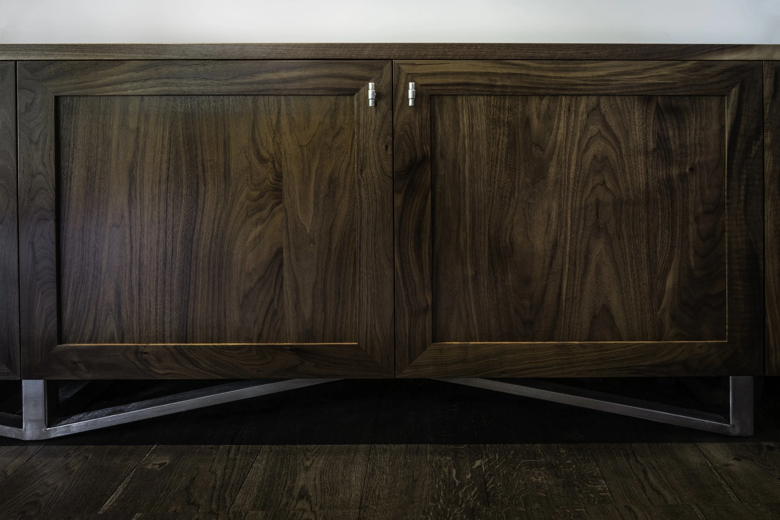 Close-up of a wooden cabinet with two doors, silver hinges, and a geometric metal base, on a wooden floor.