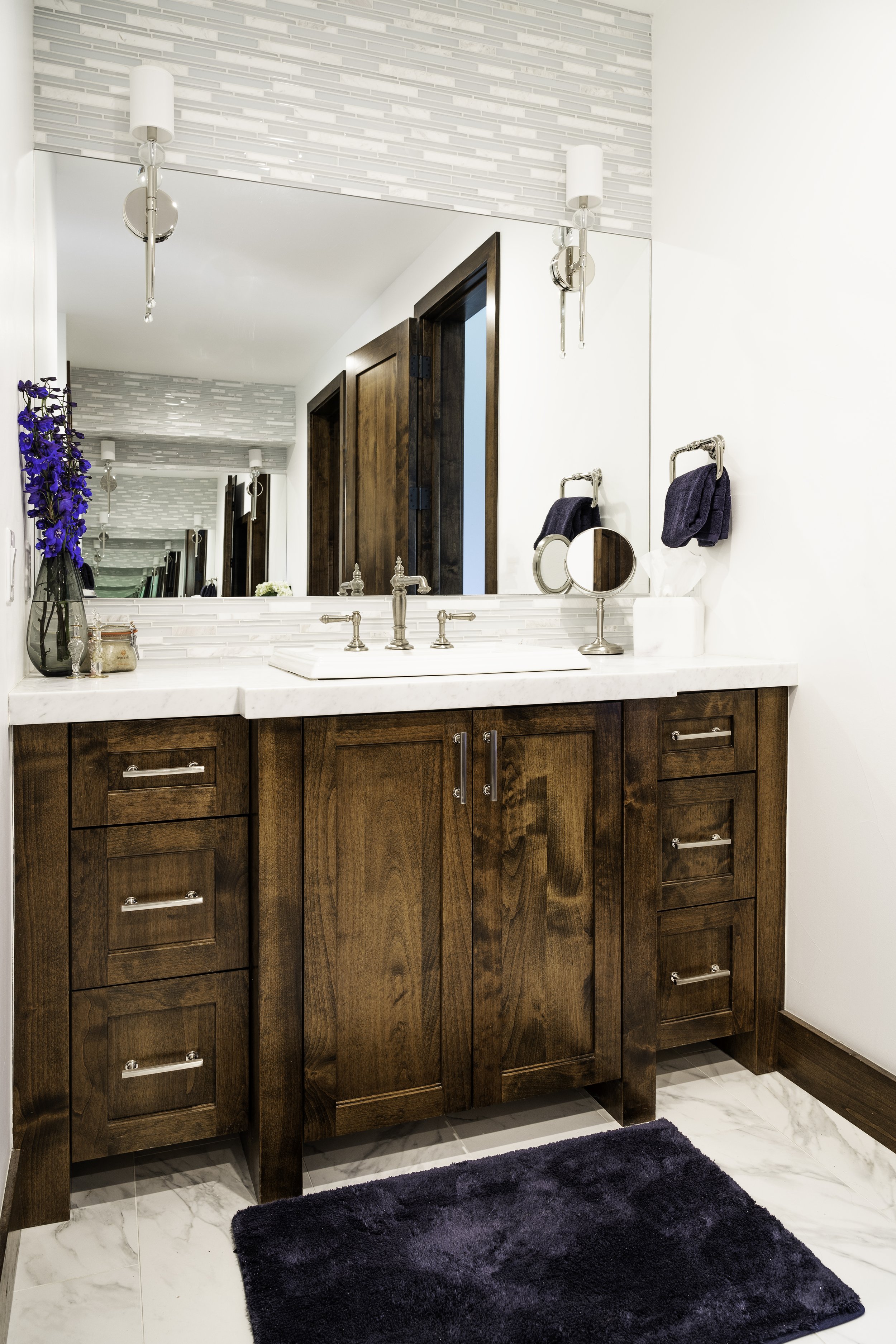 Bathroom vanity with a large mirror, white marble countertop, and wooden cabinets. Decor includes a dark blue towel, tissue box, small mirror, and a vase with blue flowers. Tiled white walls and ceiling lights.