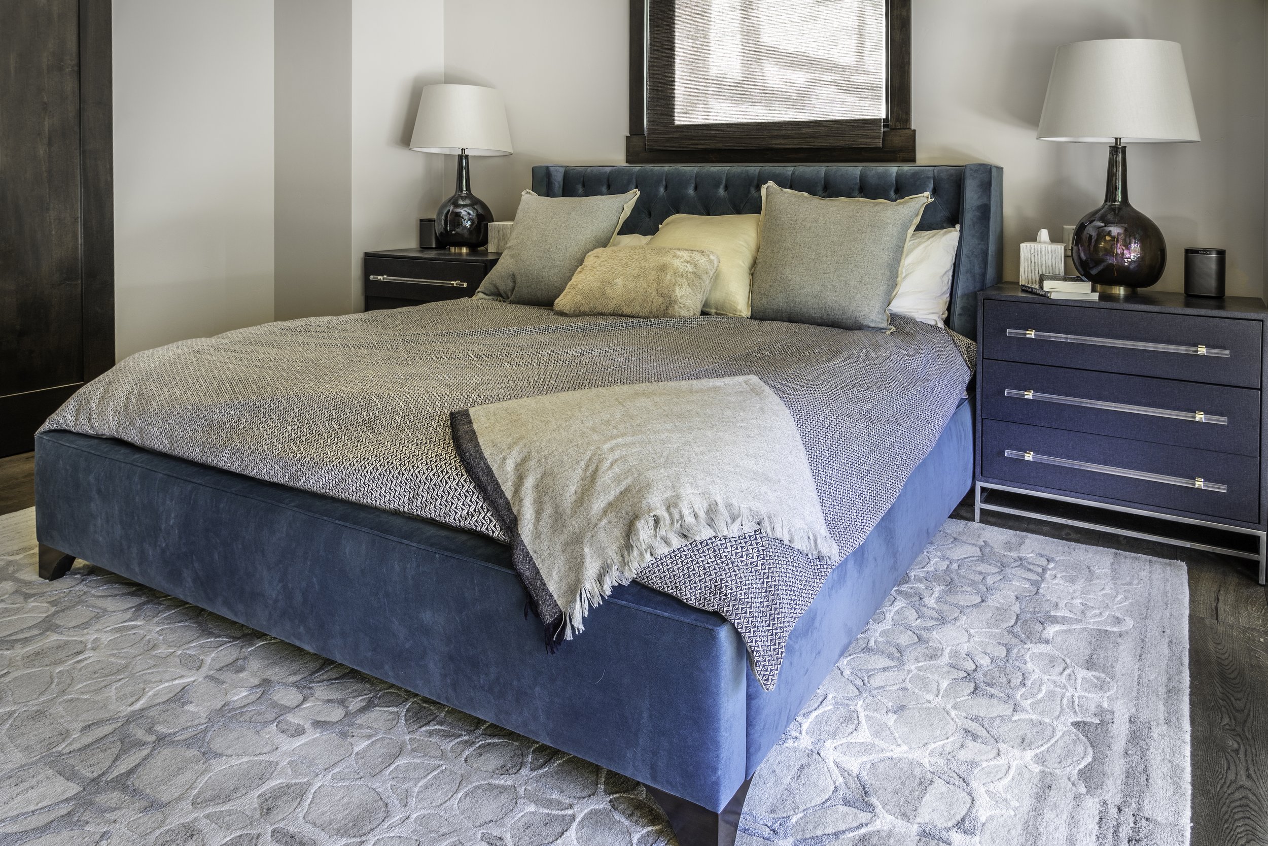 A bedroom with a large bed featuring a teal upholstered headboard and a patterned bedspread. The bed has beige, green, and textured pillows. There are two matching dark blue nightstands, each with a decorative black lamp, books, and a speaker. The ro