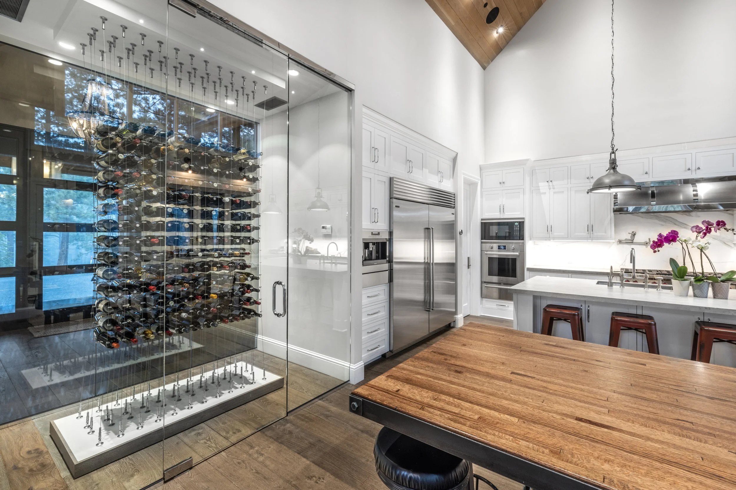 Modern kitchen with white cabinets, stainless steel appliances, and a wooden table. A glass-enclosed wine cellar with a mounted wine rack is visible, and there are three purple orchids on the counter.