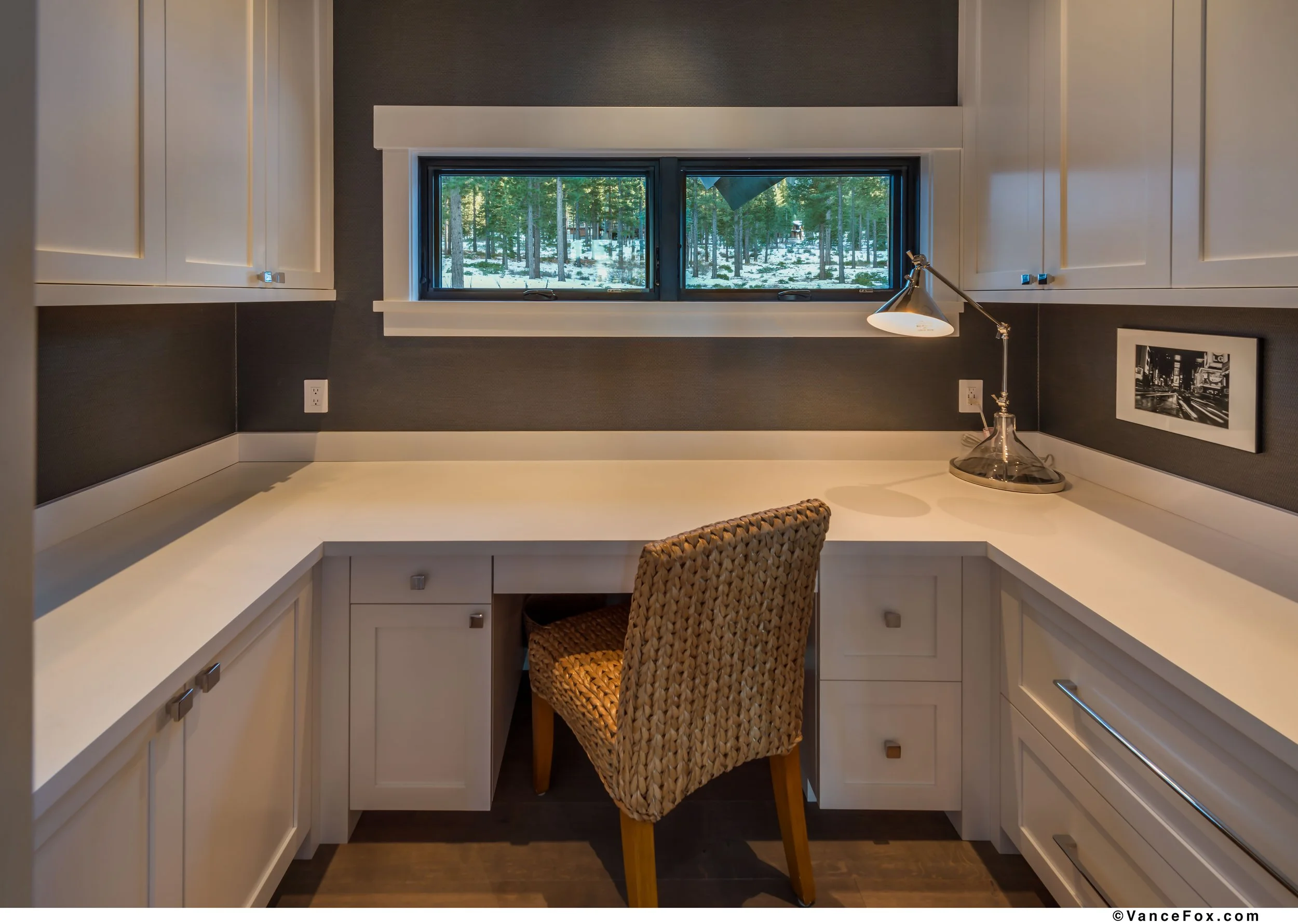 A small workspace with a white desk, beige woven chair, and a window overlooking a snowy forest. There is a desk lamp and a black-and-white framed photograph on the wall.