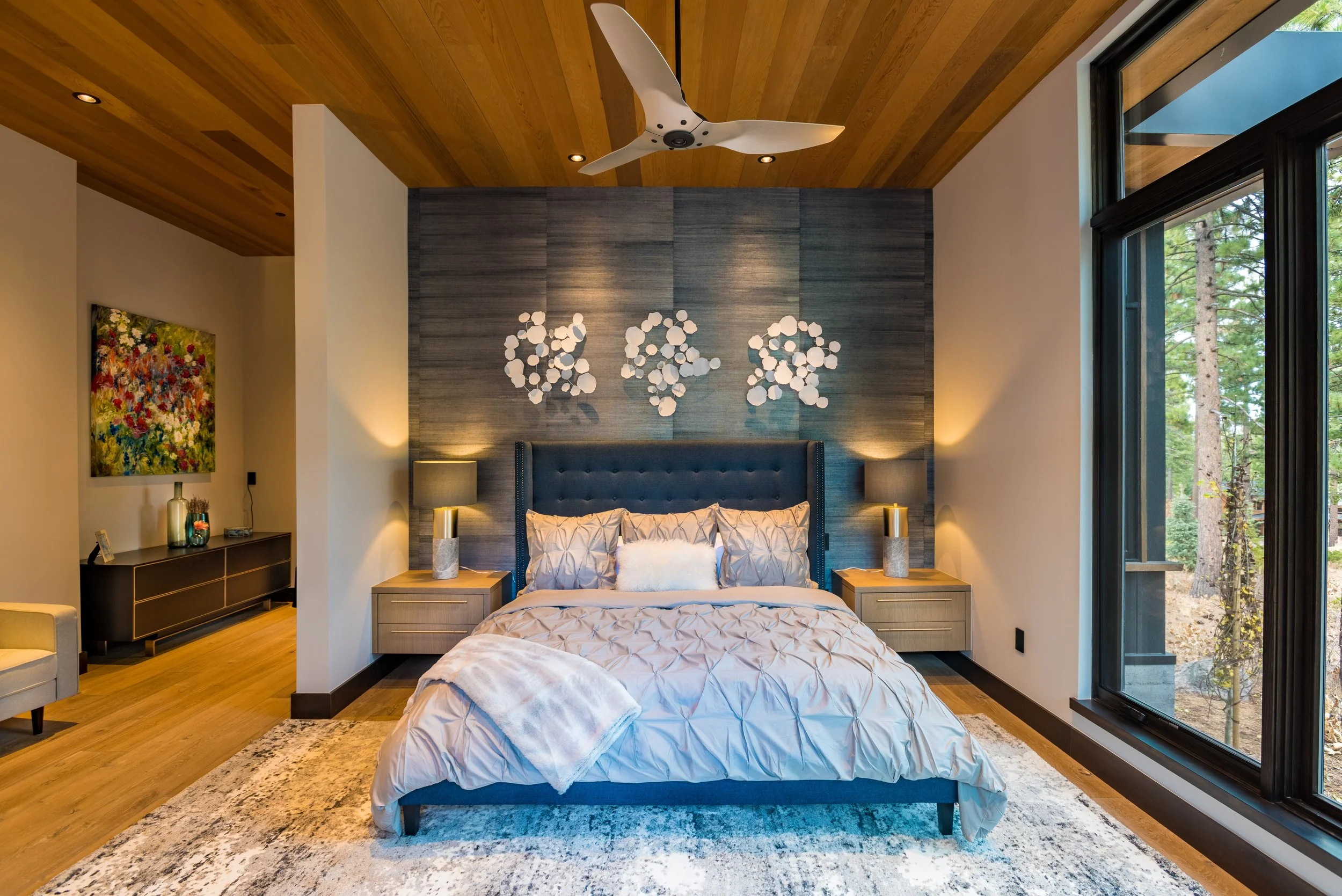 Modern bedroom with large bed, blue headboard, two nightstands with lamps, large window showing trees outside, wood ceiling, artwork on wall, ceiling fan, and a rug.