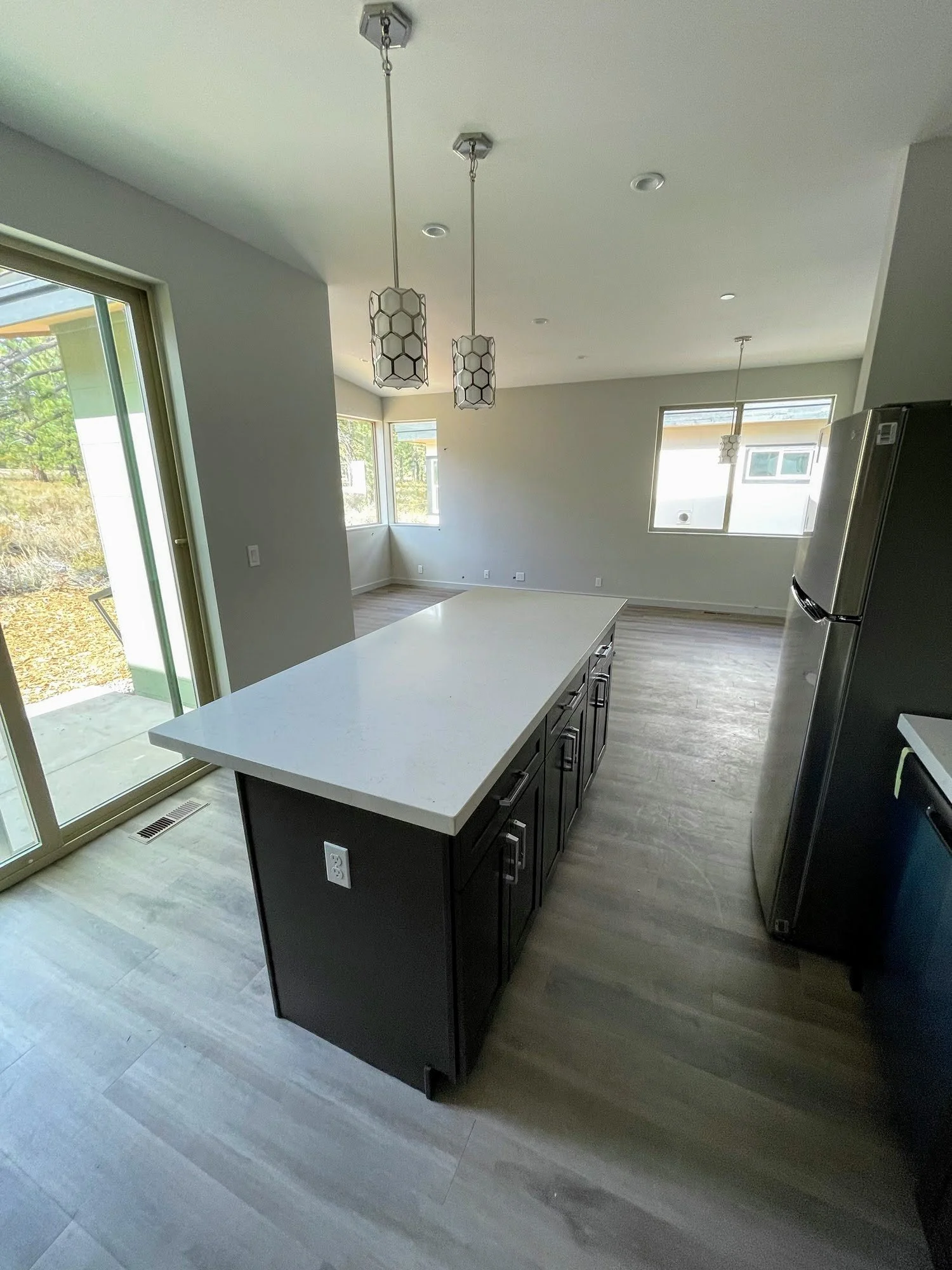 Modern kitchen with a white countertop island, black cabinets, stainless steel refrigerator, pendant lights, and large windows letting in natural light.