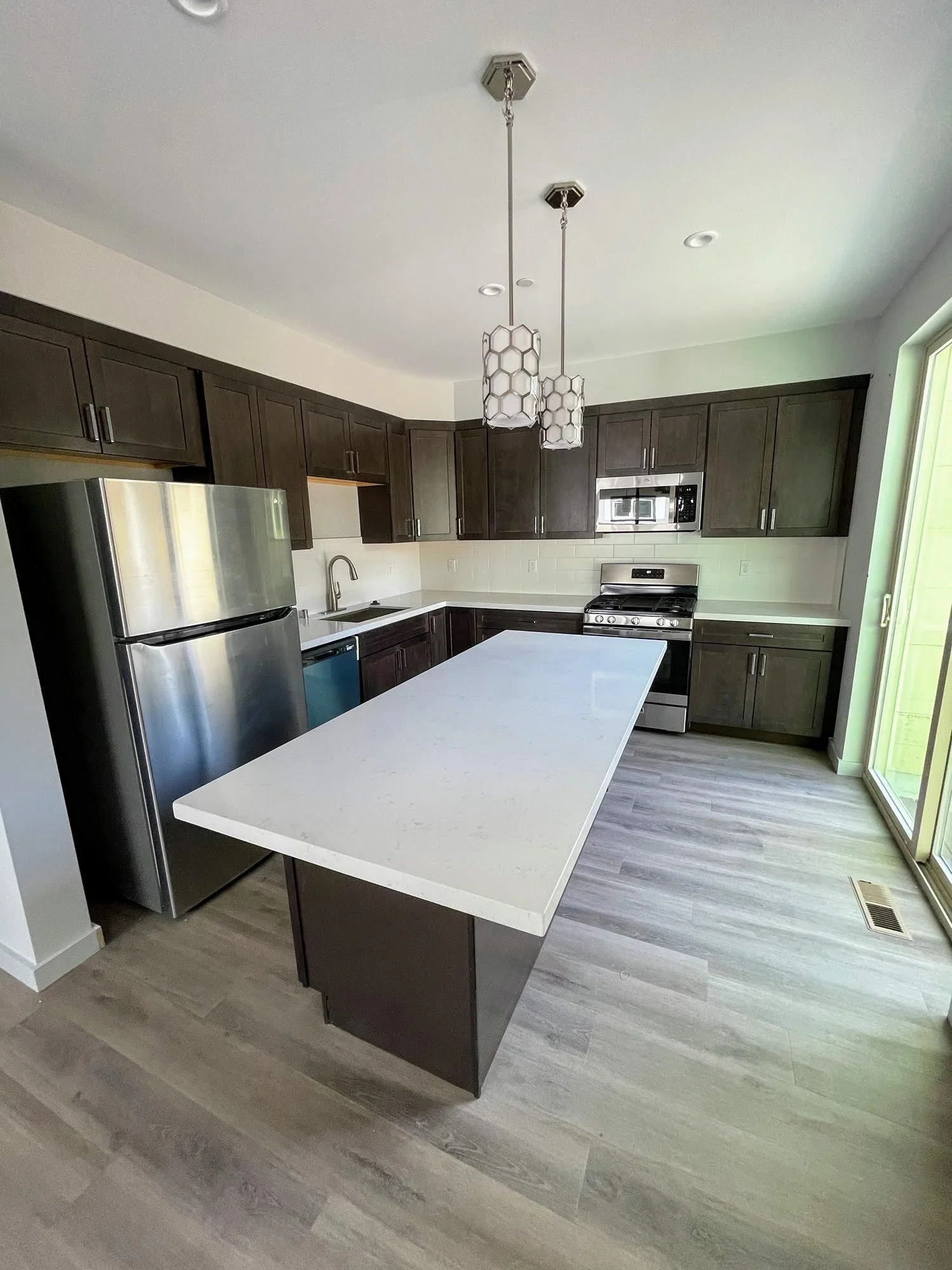 Modern kitchen with dark wood cabinets, stainless steel appliances, a large white island, and pendant lights with a honeycomb pattern.