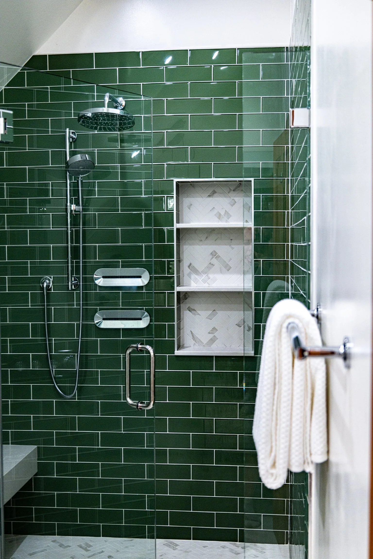 Green tiled shower with glass door, built-in shelves, and shower fixtures including a rain shower head and a handheld shower.