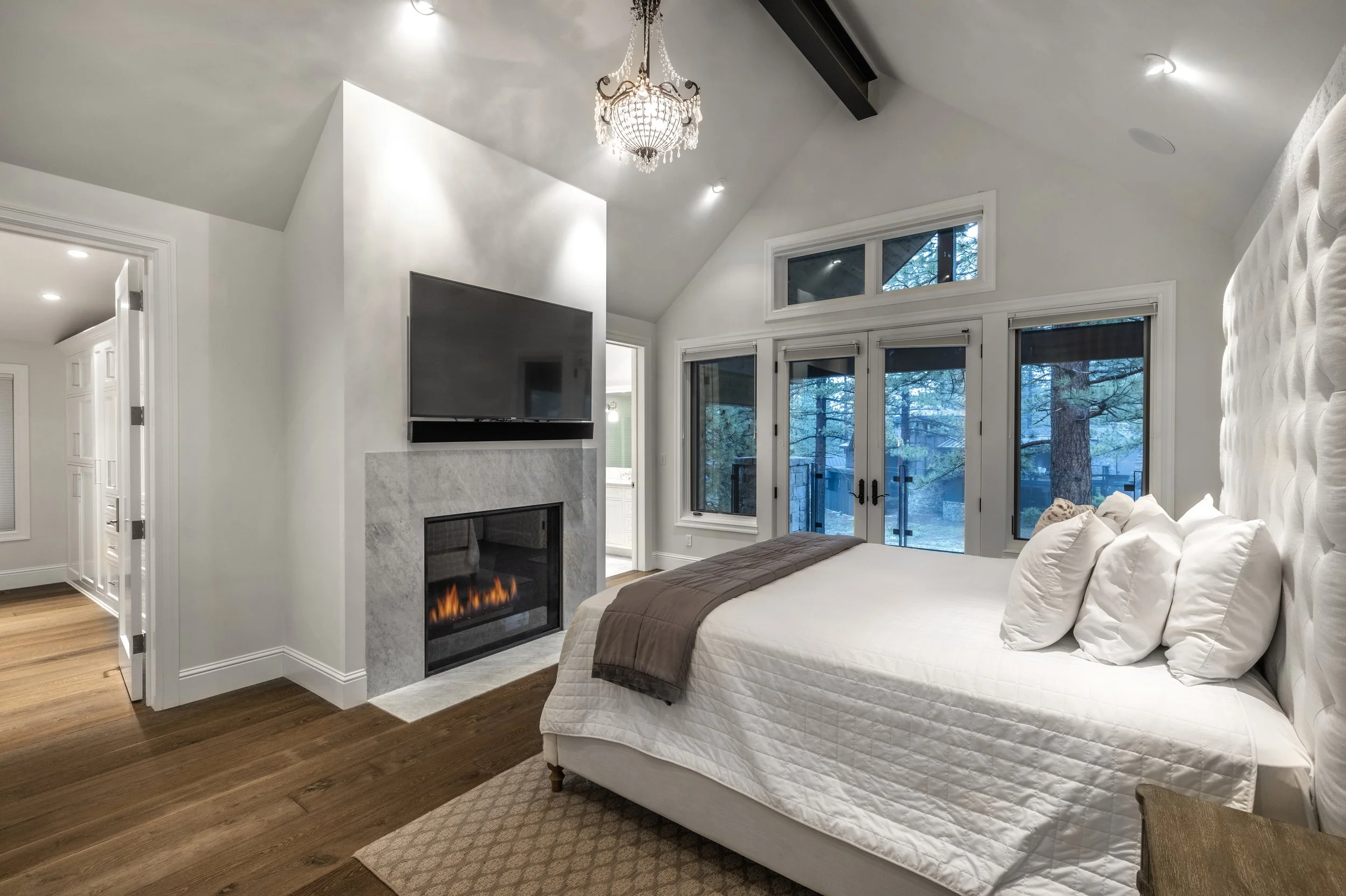 Modern bedroom with large sliding glass doors leading to a wooded outdoor area, a white bed with multiple pillows, a brown throw, a fireplace with a mounted TV above, and a chandelier hanging from the ceiling.