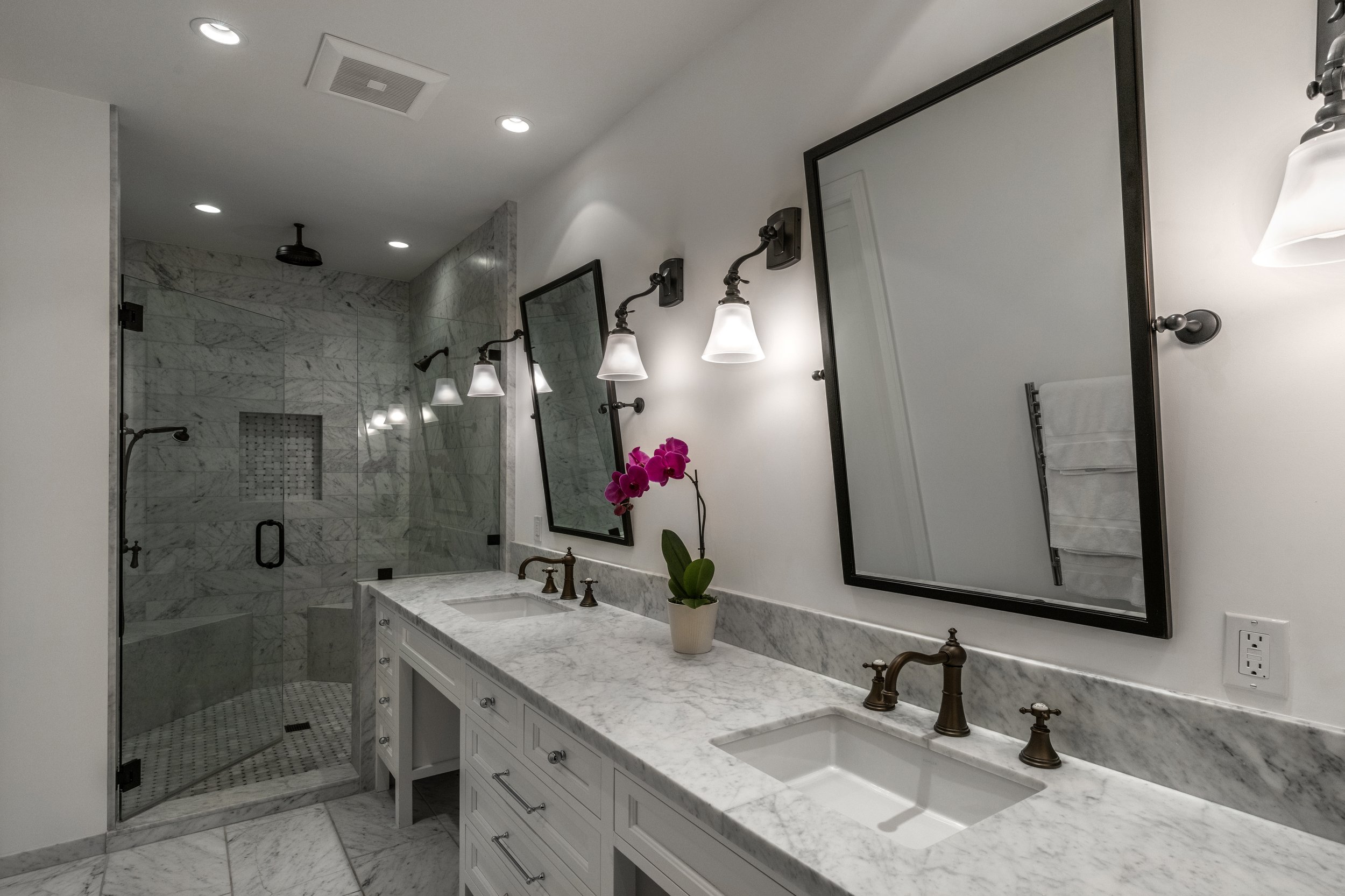 Modern bathroom with double vanity, marble countertop, two rectangular mirrors with black frames, wall-mounted light fixtures, a potted purple orchid, and a glass-enclosed marble shower in the background.