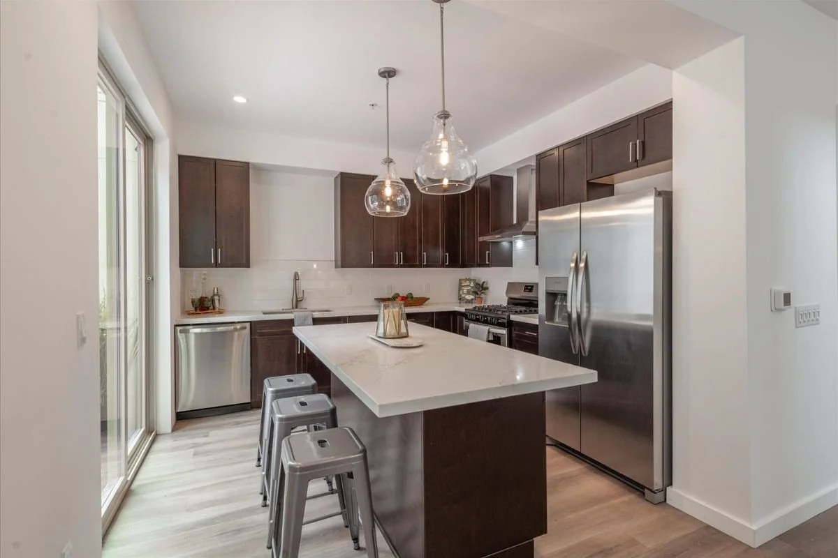 Modern kitchen with dark wooden cabinets, stainless steel refrigerator, dishwasher, and stove, white countertops, and pendant lights over island with three metal stools.