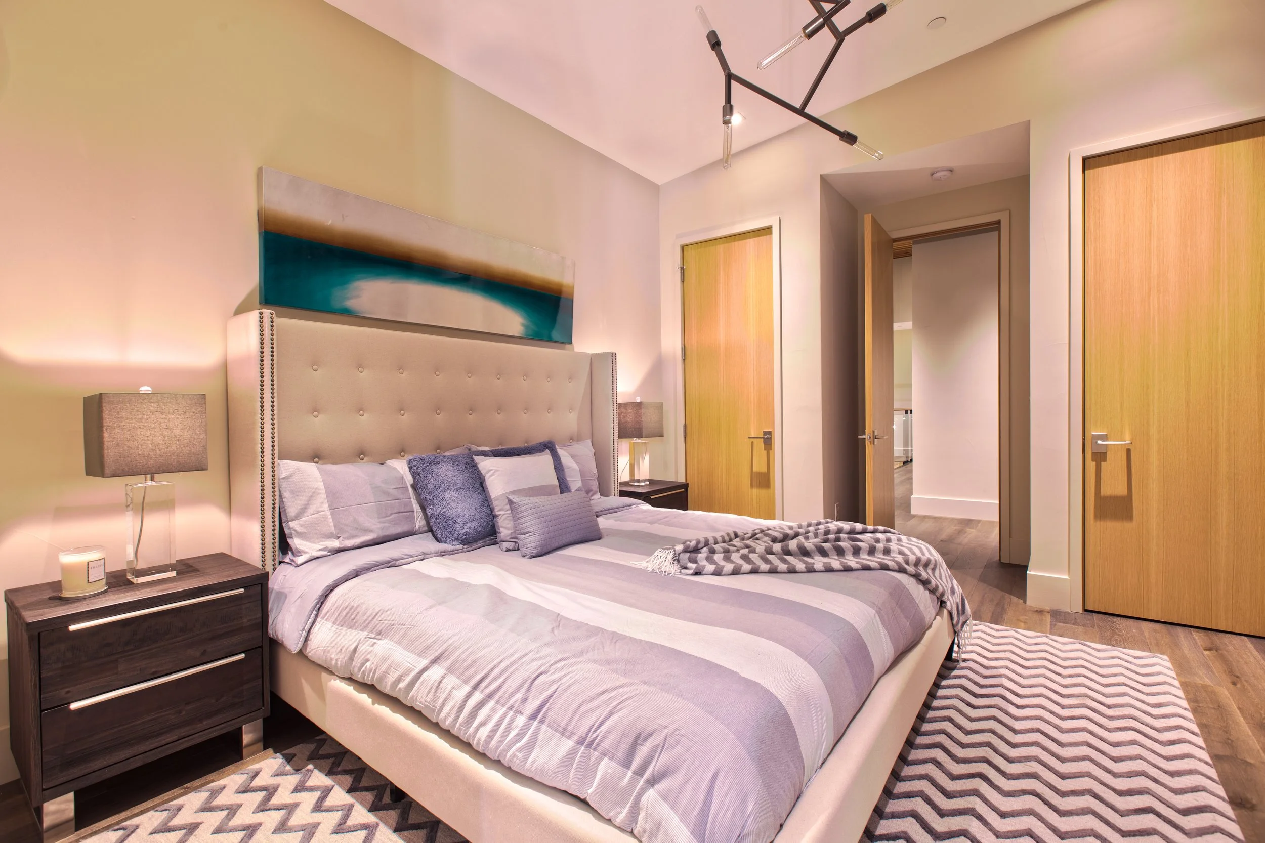 Modern bedroom with a large upholstered bed, striped bedding, gray pillows, and a black nightstand with a lamp, candle, and decor. Artwork hangs above the headboard, and three wooden doors are visible, with a Chevron-patterned rug on wooden floors.