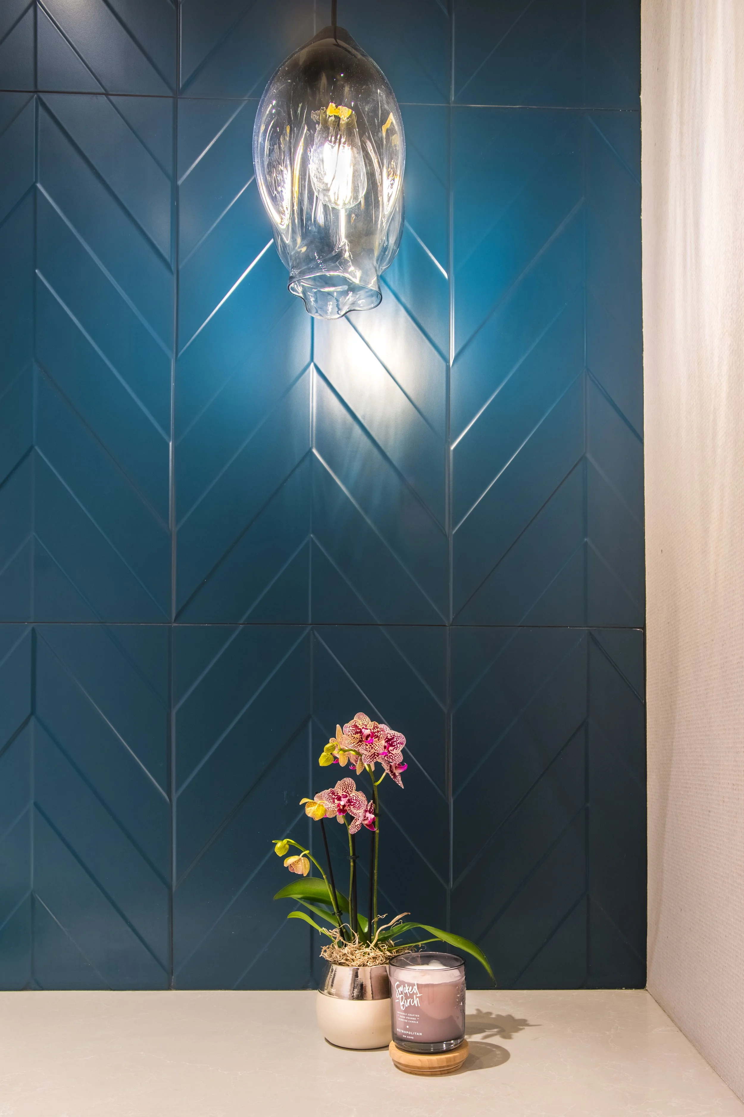 A potted orchid plant with pink and white flowers, a candle in a glass container, and a modern glass wall sconce on a beige surface against a dark blue tiled wall.