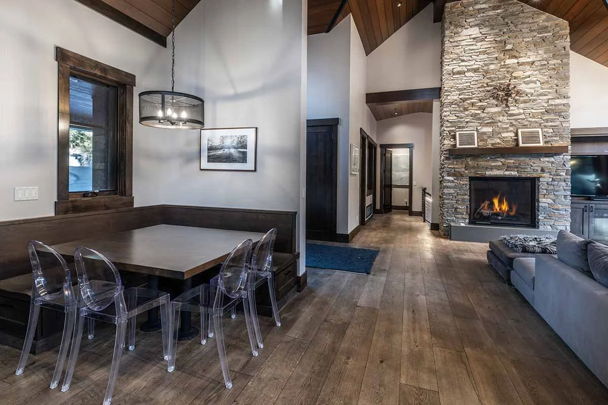 A living room and dining area with wood flooring, a stone fireplace, a gray sofa, a wooden dining table with transparent chairs, a window with wooden trim, and a modern light fixture hanging above the dining area.