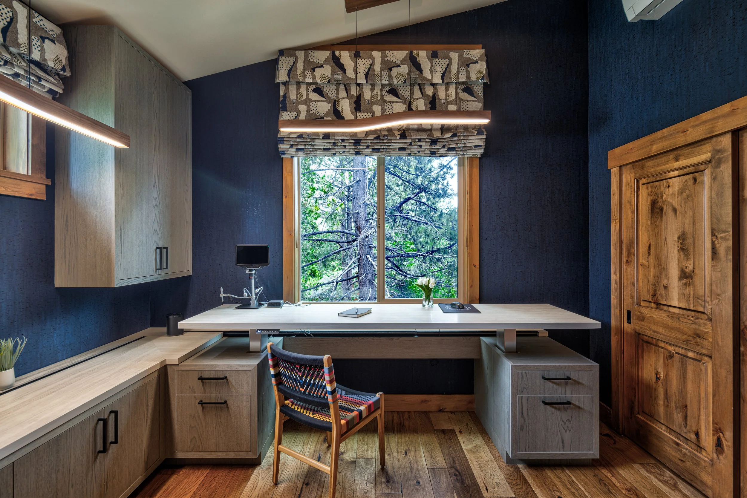 Home office with a large desk facing a window with trees outside, dark blue walls, wood accents, and a colorful chair.