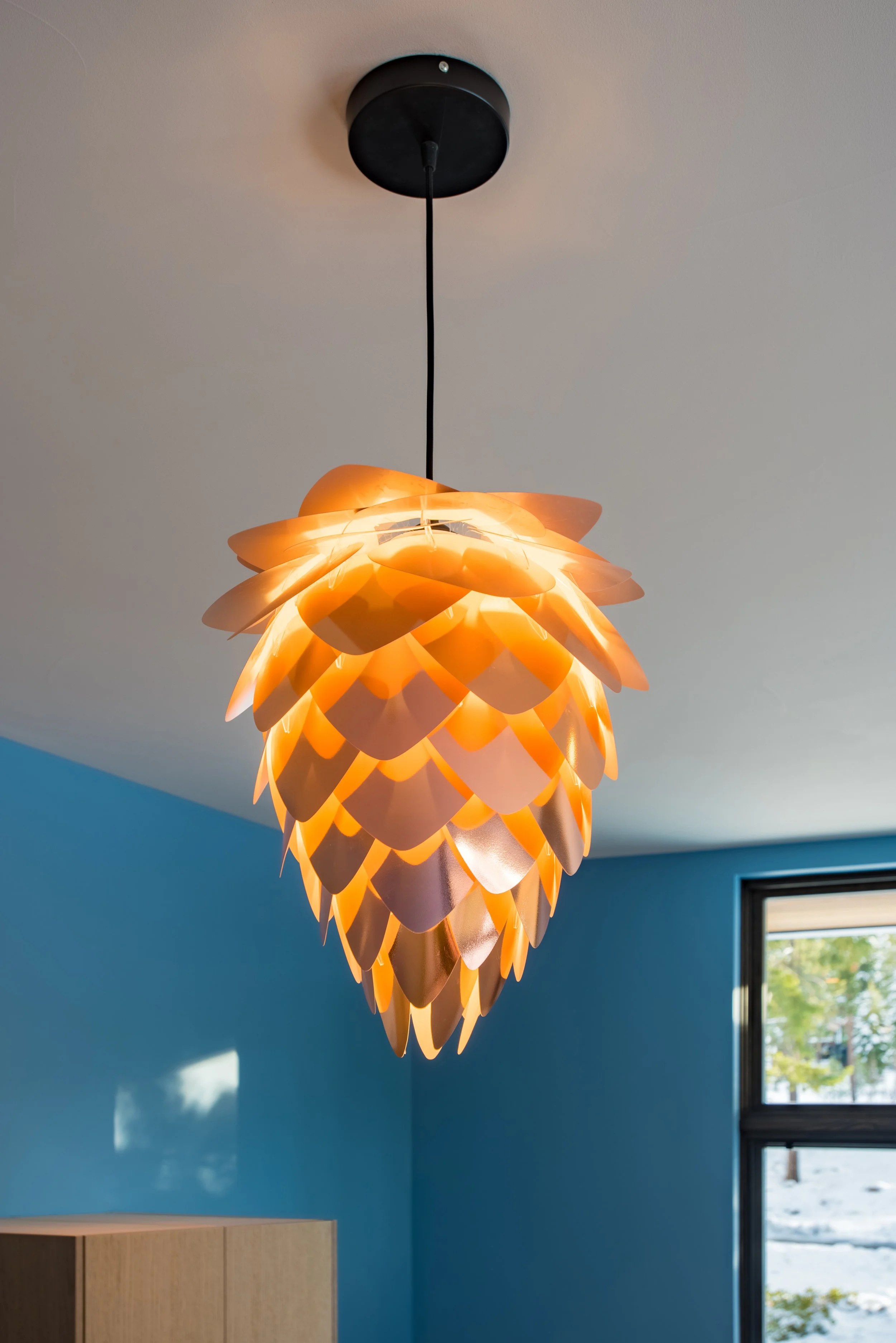 Modern pendant ceiling light with petal-like orange and pink shades hanging over a room with a blue wall and a window showing outdoor trees.