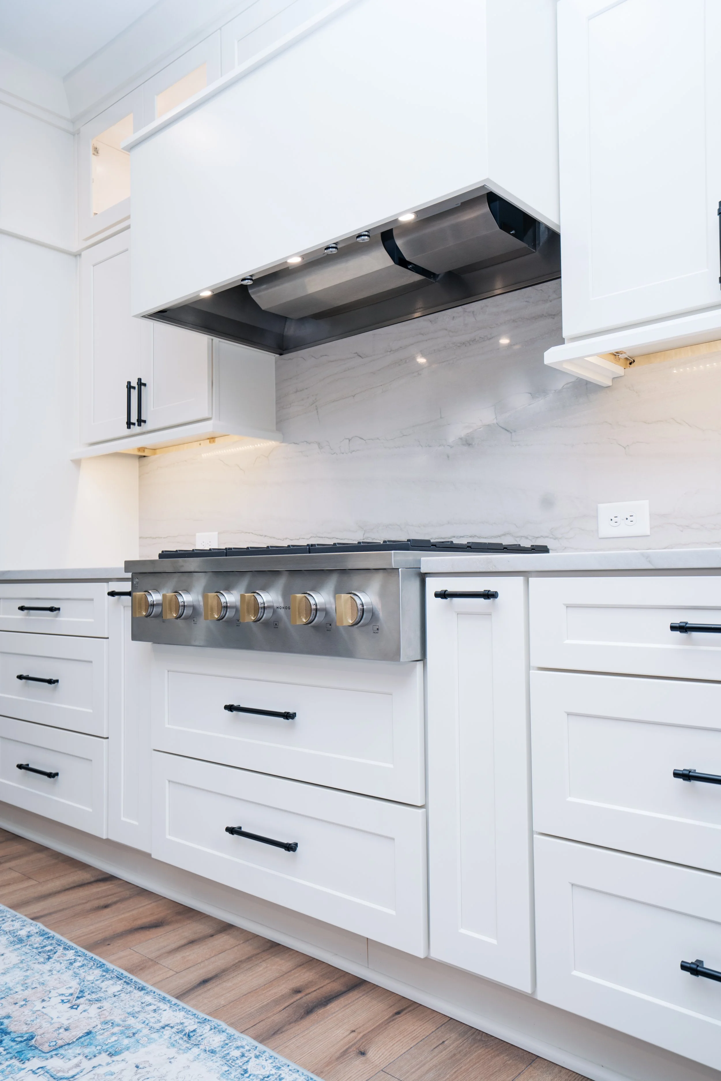 Modern white kitchen with black cabinet handles, stainless steel stove with gold knobs, white marble backsplash, and wood flooring.