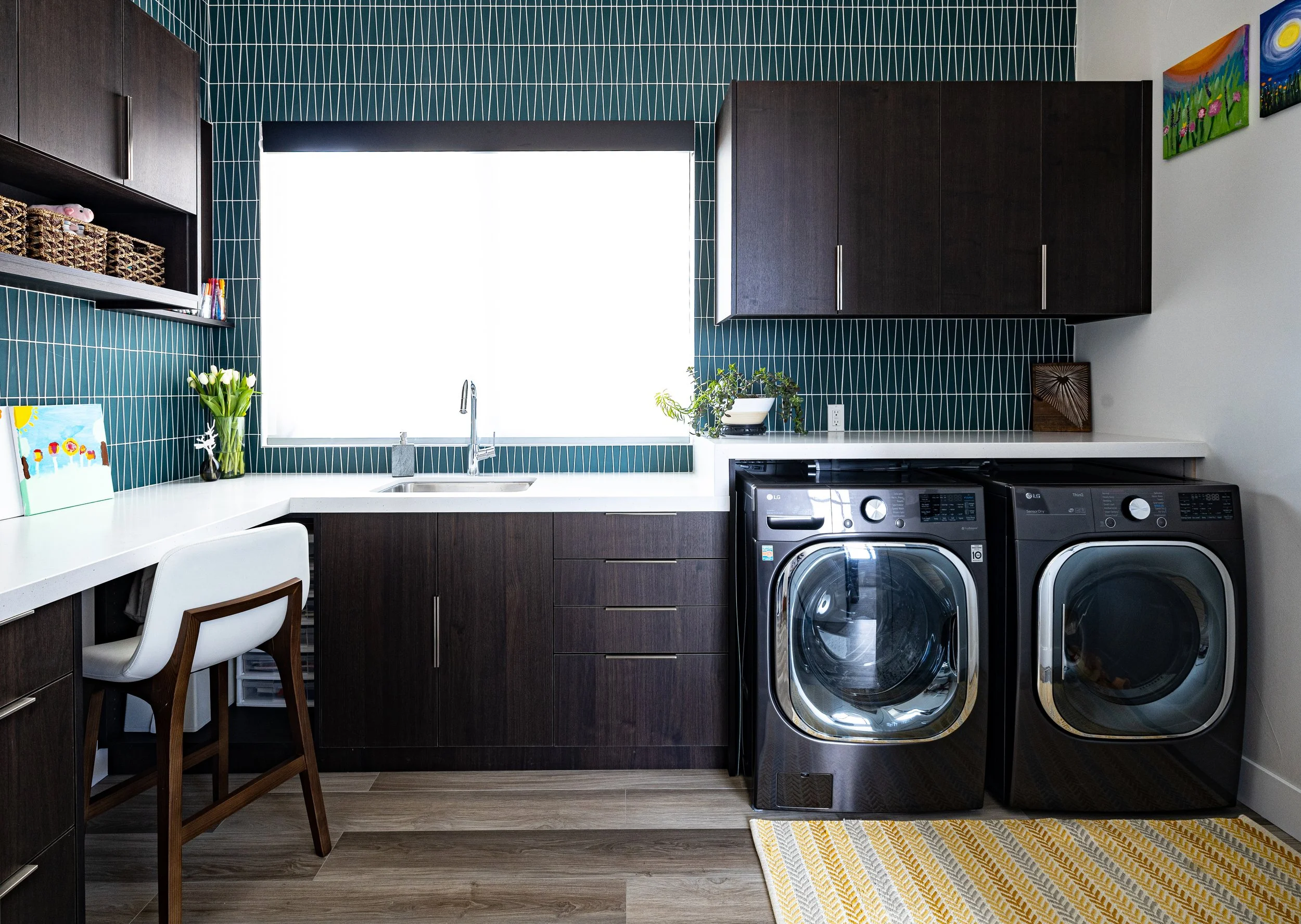Laundry room with dark wood cabinets, a white countertop, a window with a black blind, front-loading washer and dryer, decorative artwork, and indoor plants.