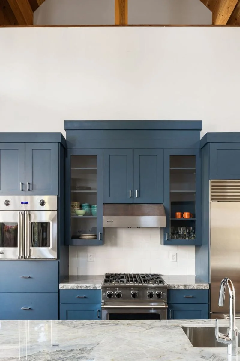A modern kitchen with blue cabinets, a stainless steel oven and refrigerator, open shelves with dishes, and a marble countertop island.