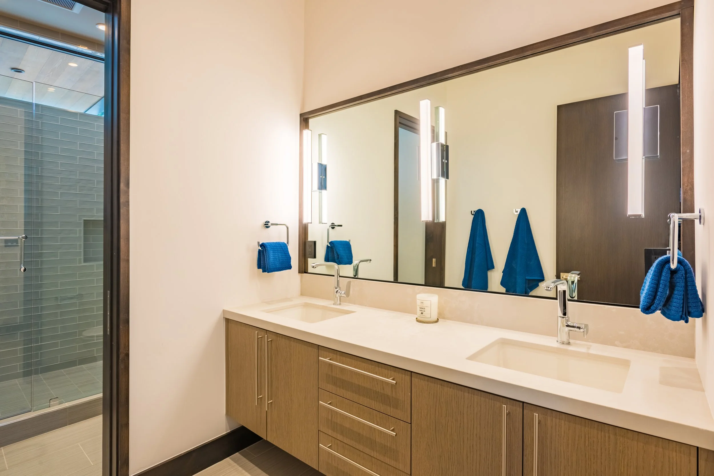 Modern bathroom with double sink vanity, large mirror, and blue towels, adjacent to a walk-in glass shower with gray tiles.