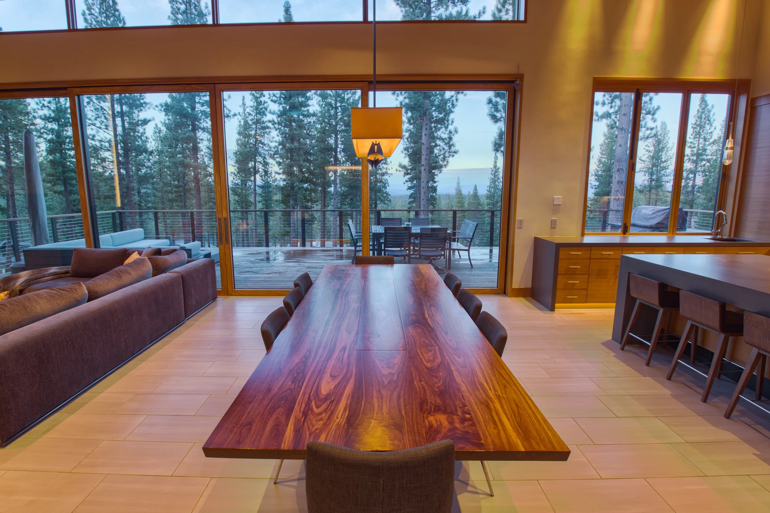 Interior of a modern house with large glass sliding doors leading to a wooded outdoor deck with outdoor furniture, followed by a dining area with a long wooden table and chairs, a purple sectional sofa, and a kitchen window with a view of trees.