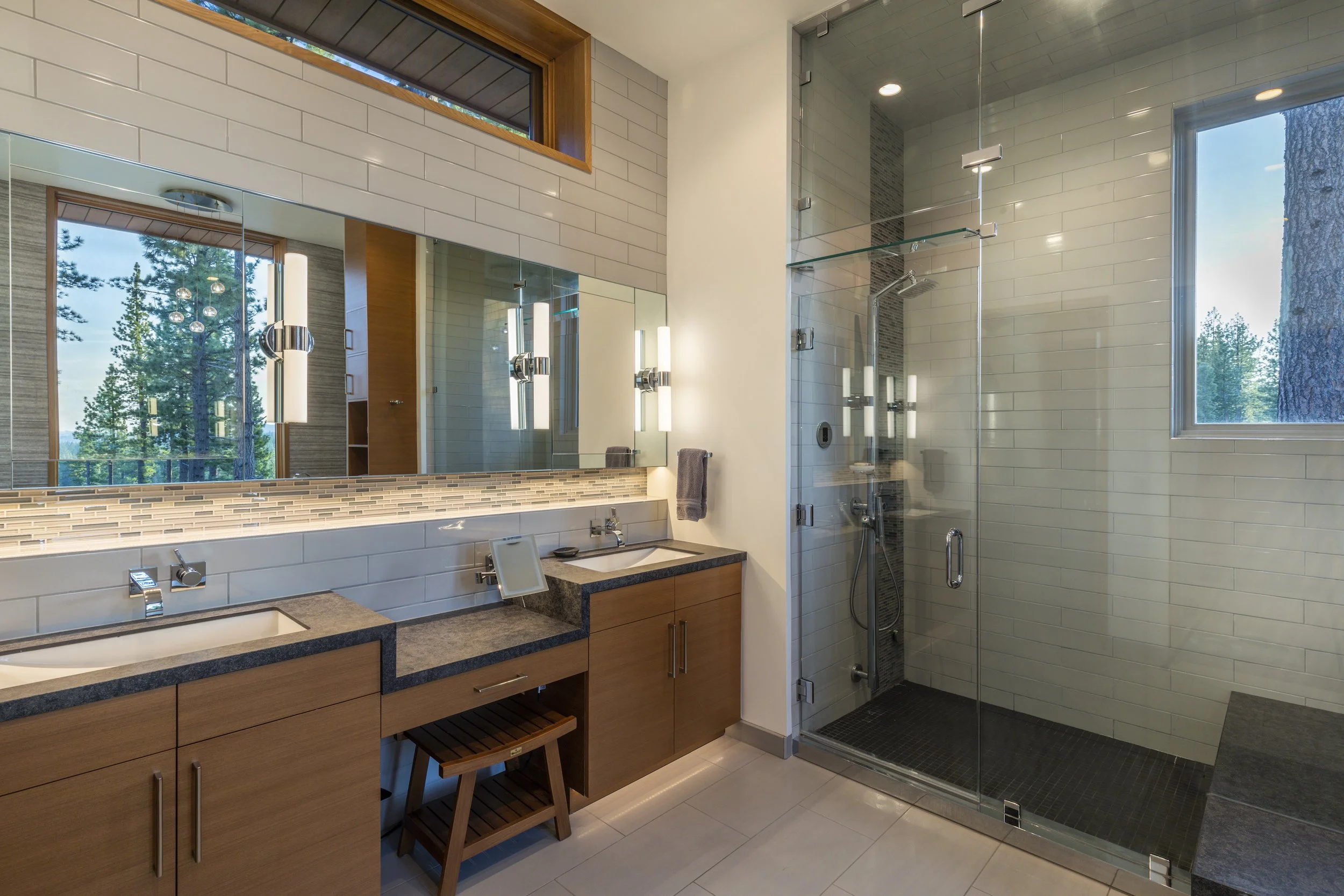 Modern bathroom with dual sinks, large mirror, and glass shower enclosure in a well-lit space with a view of trees outside.