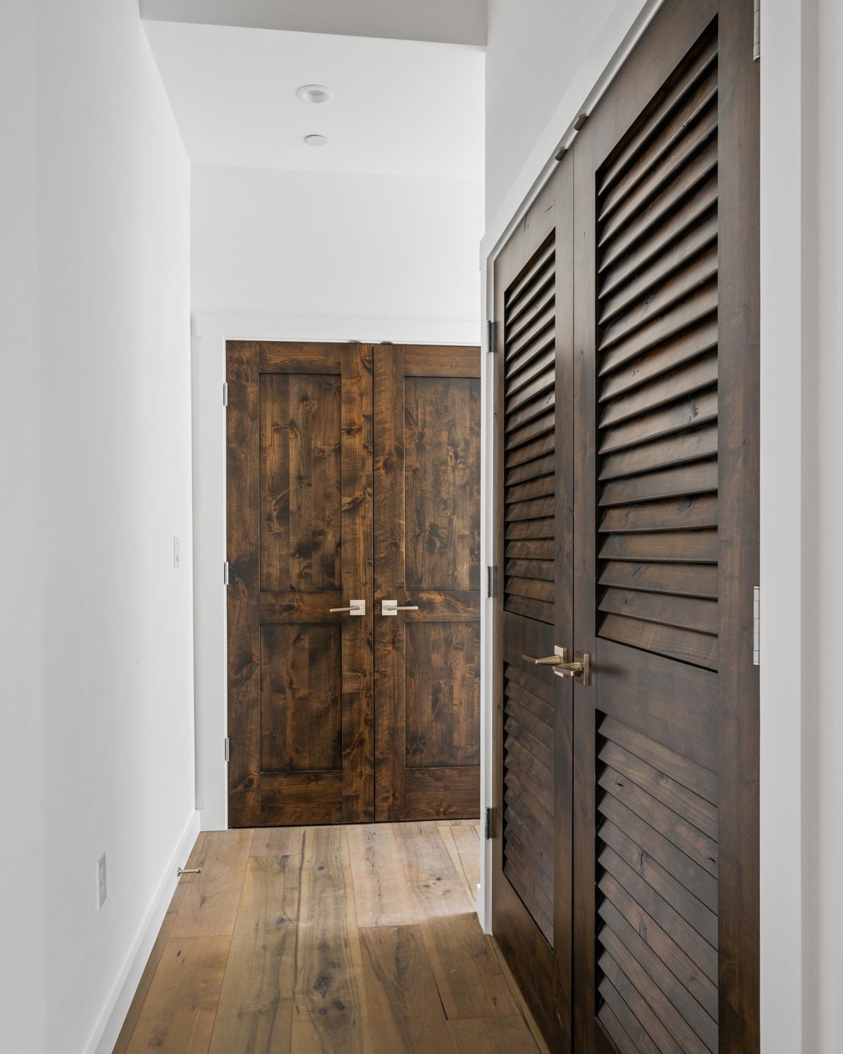 Hallway with white walls, hardwood floor, and dark wood double doors, with a wooden slatted closet door on the right.