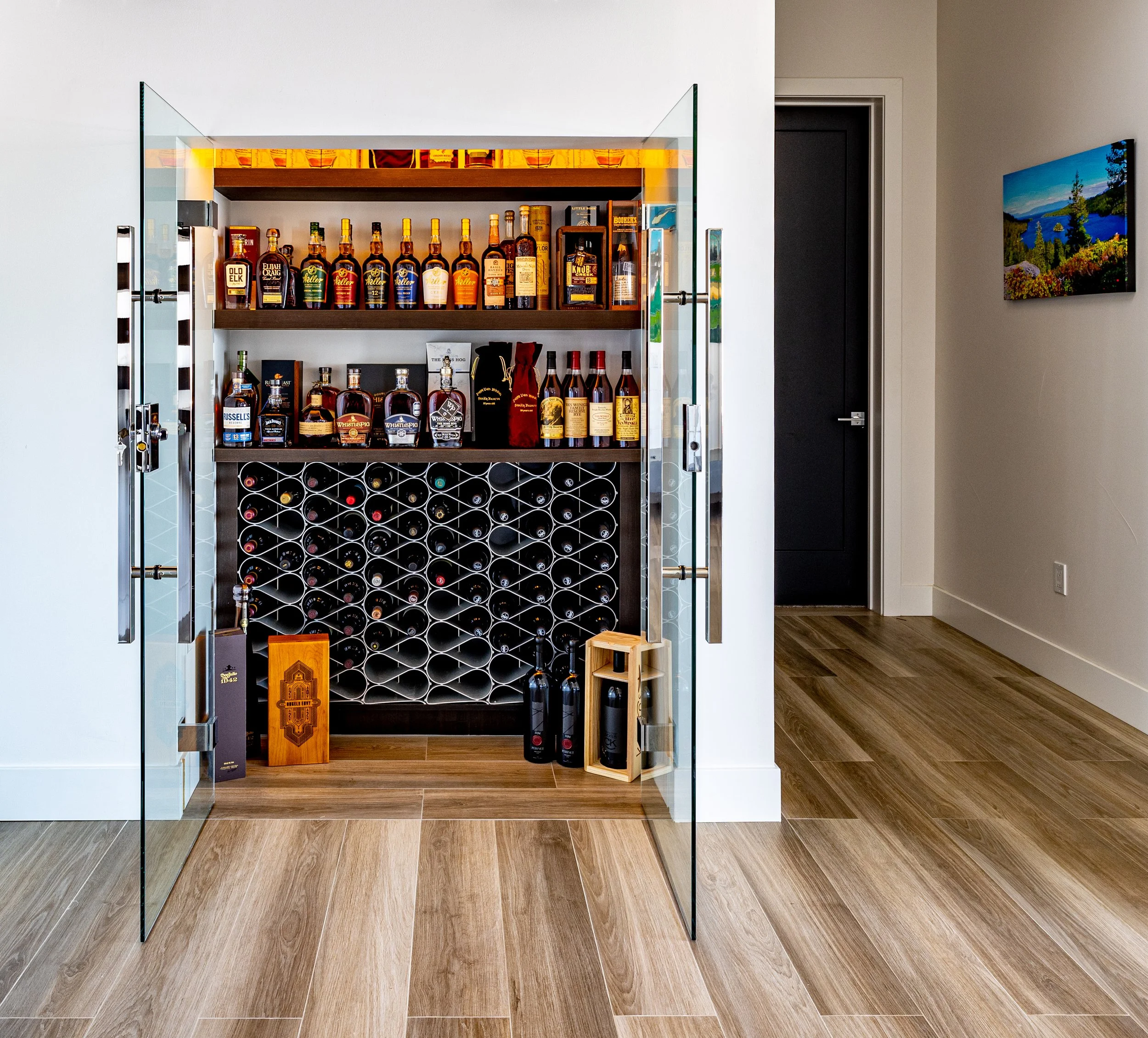 A glass door wine cellar with shelves of liquor bottles, wine bottles, and wine racks inside, situated in a room with wooden floor and a white wall, with a doorway to the right and a framed landscape picture on the wall.