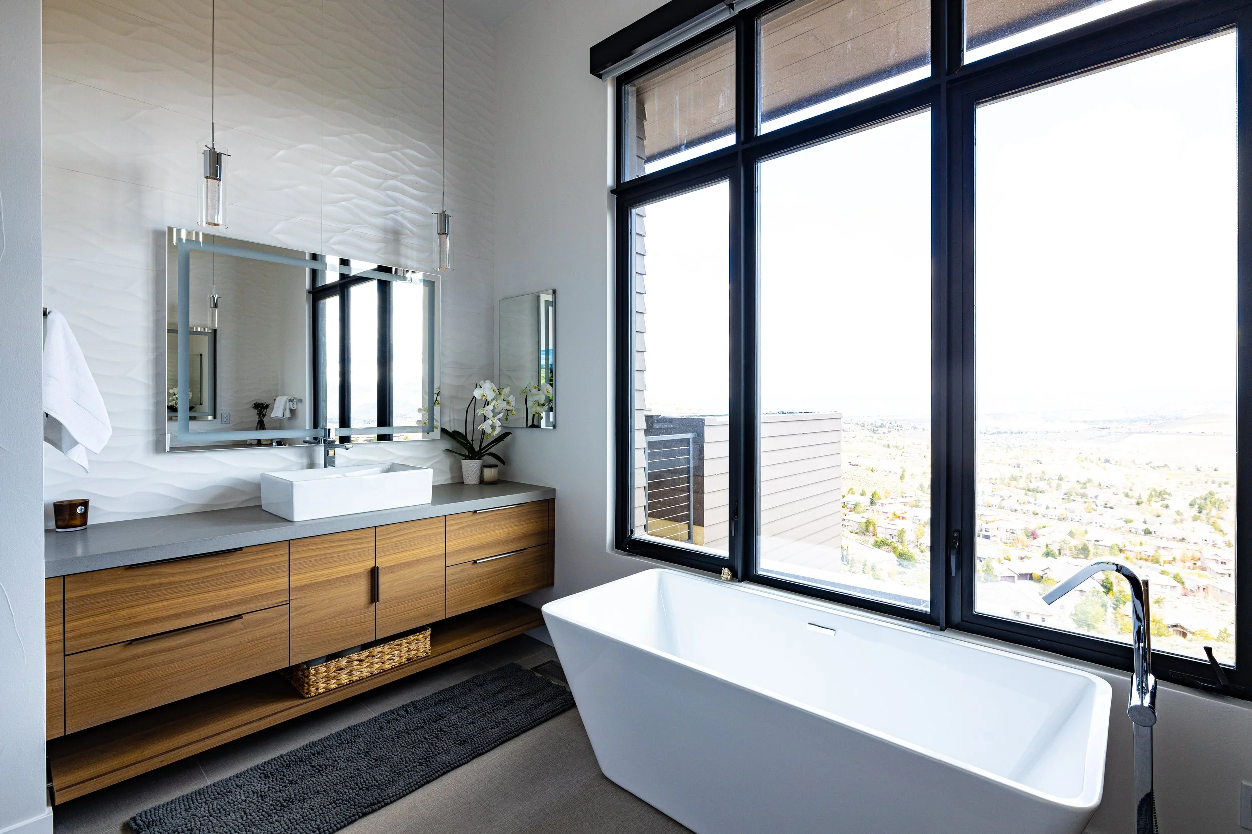 Modern bathroom with a large freestanding bathtub near a window with a scenic view, a wooden vanity with a stone countertop, a rectangular vessel sink, a mirror, and decorative flowers.
