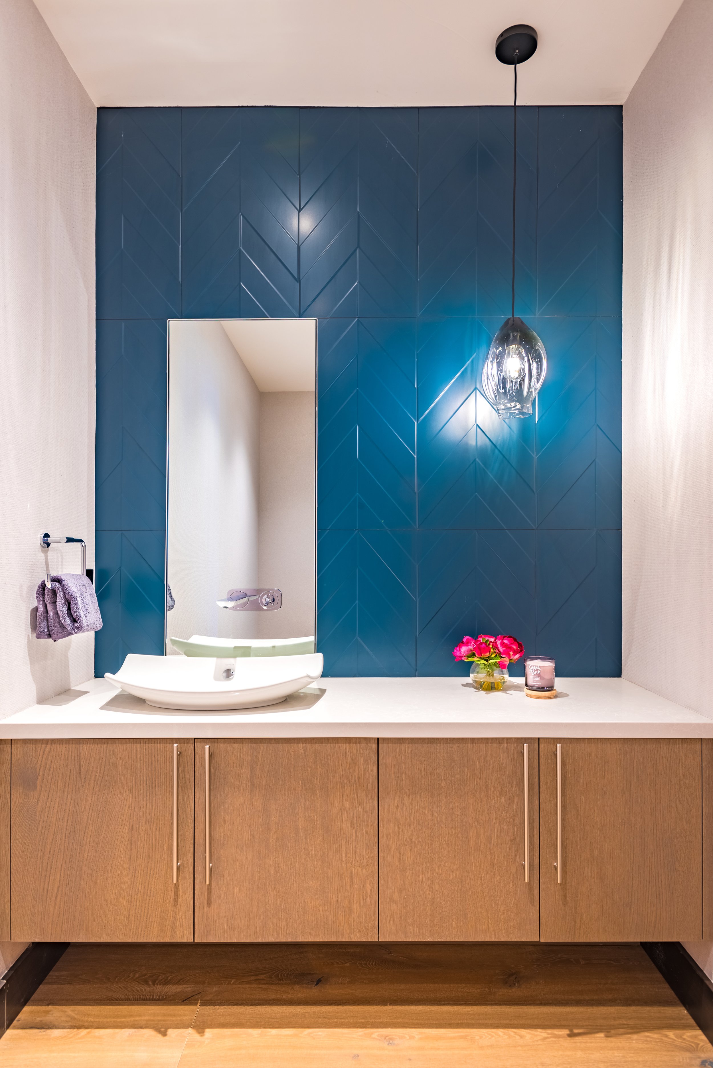 Bathroom vanity with a wooden cabinet, a white countertop, a vessel sink, a mirror, purple towels, decorative flowers, a candle, and a hanging light fixture against a blue tiled wall.