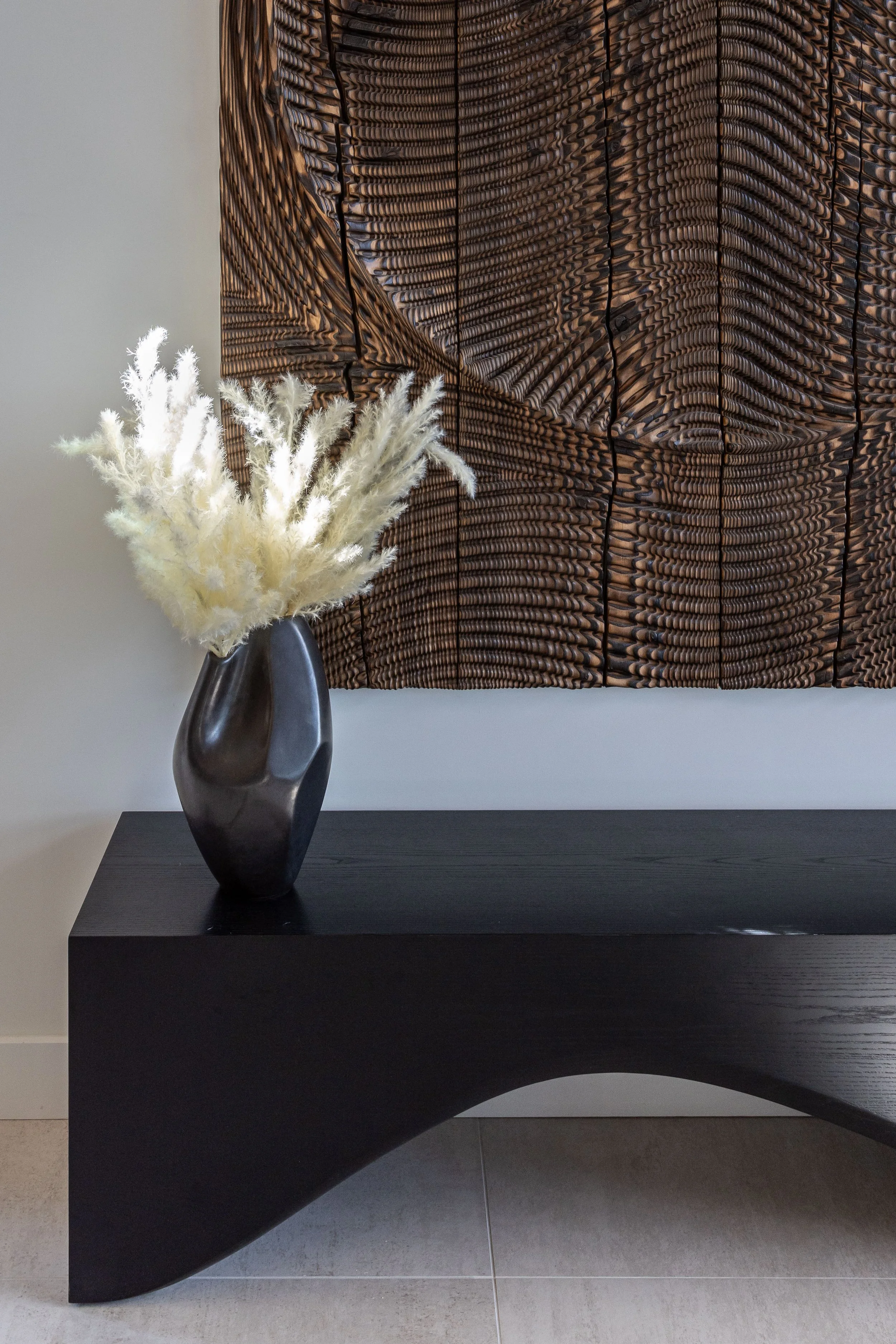 A dark, modern wooden table with a black, glossy vase holding white pampas grass foliage, positioned in front of a textured brown and black abstract wall hanging.