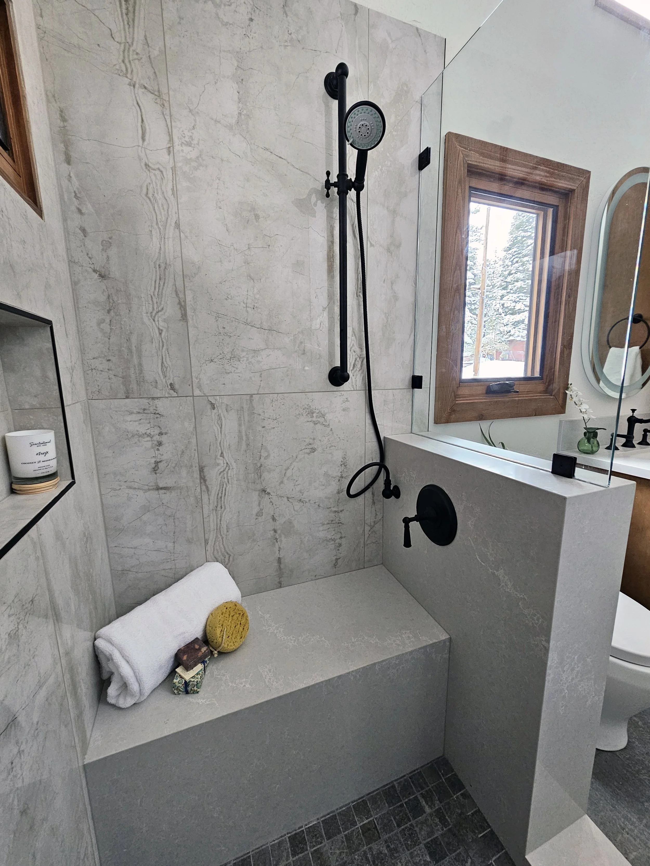A modern bathroom shower with a black rainfall showerhead and handheld shower on a black bar, beige marble walls, a window with a wooden frame, a white towel, spa accessories, and a partial view of a toilet and mirror.