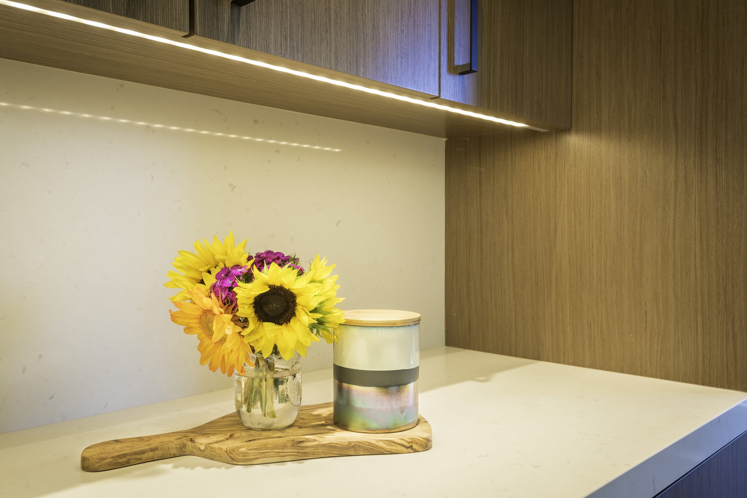 A vase with a bouquet of yellow sunflowers and pink flowers on a cutting board, next to a cylindrical container with a lid on a white countertop with wooden cabinets in the background.