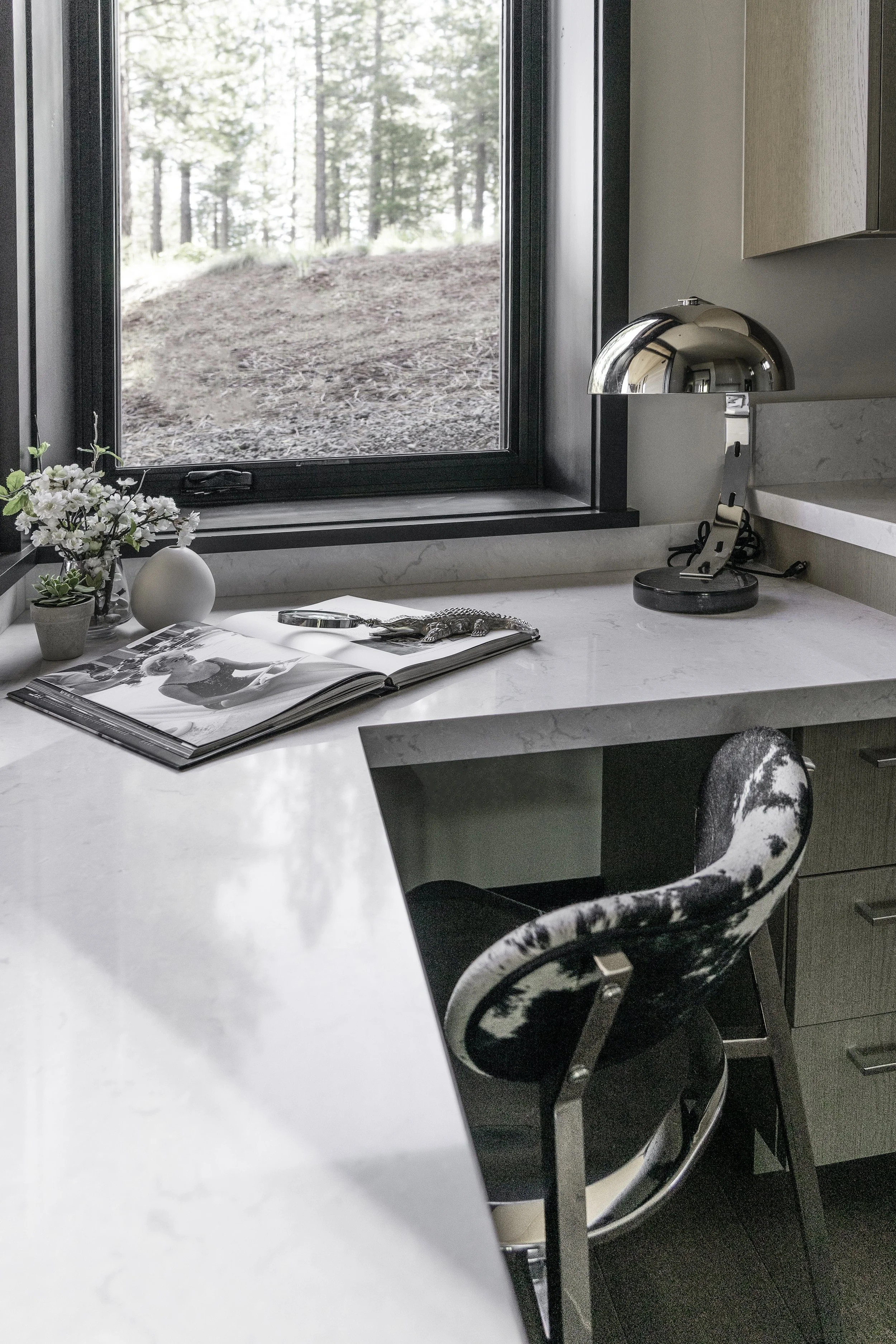 A modern kitchen with a white marble countertop, a black and white cowhide-patterned chair, a small plant, a vase, an open magazine, and a silver desk lamp near a window overlooking a wooded outdoor scene.