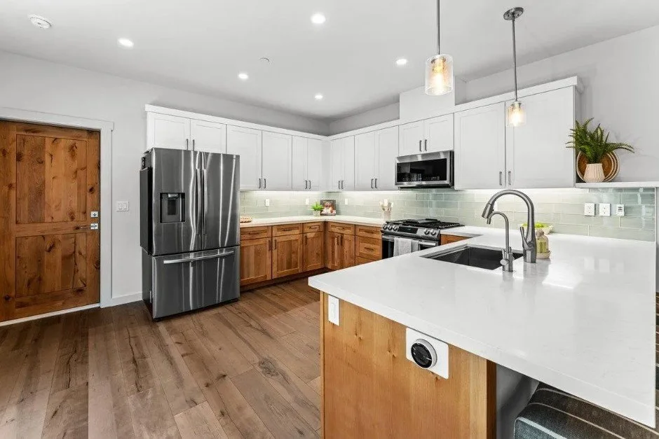 Modern kitchen with white cabinets, wooden lower cabinets, stainless steel refrigerator, microwave, gas stove, and white countertops. Includes a sink with a tall faucet, a potted plant, and pendant lights.