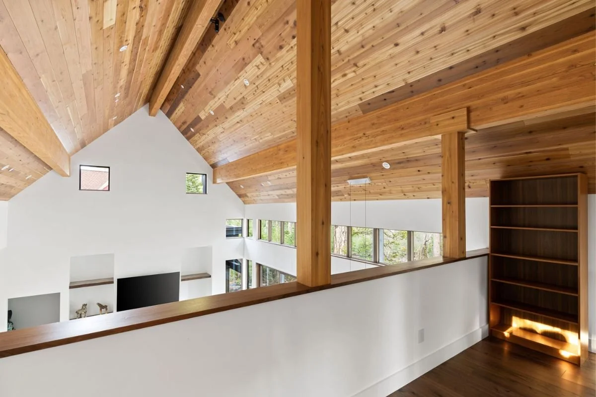 Interior view of a modern house with a high wooden ceiling, large windows, a white wall with niches, a mounted TV, and a wooden bookshelf.