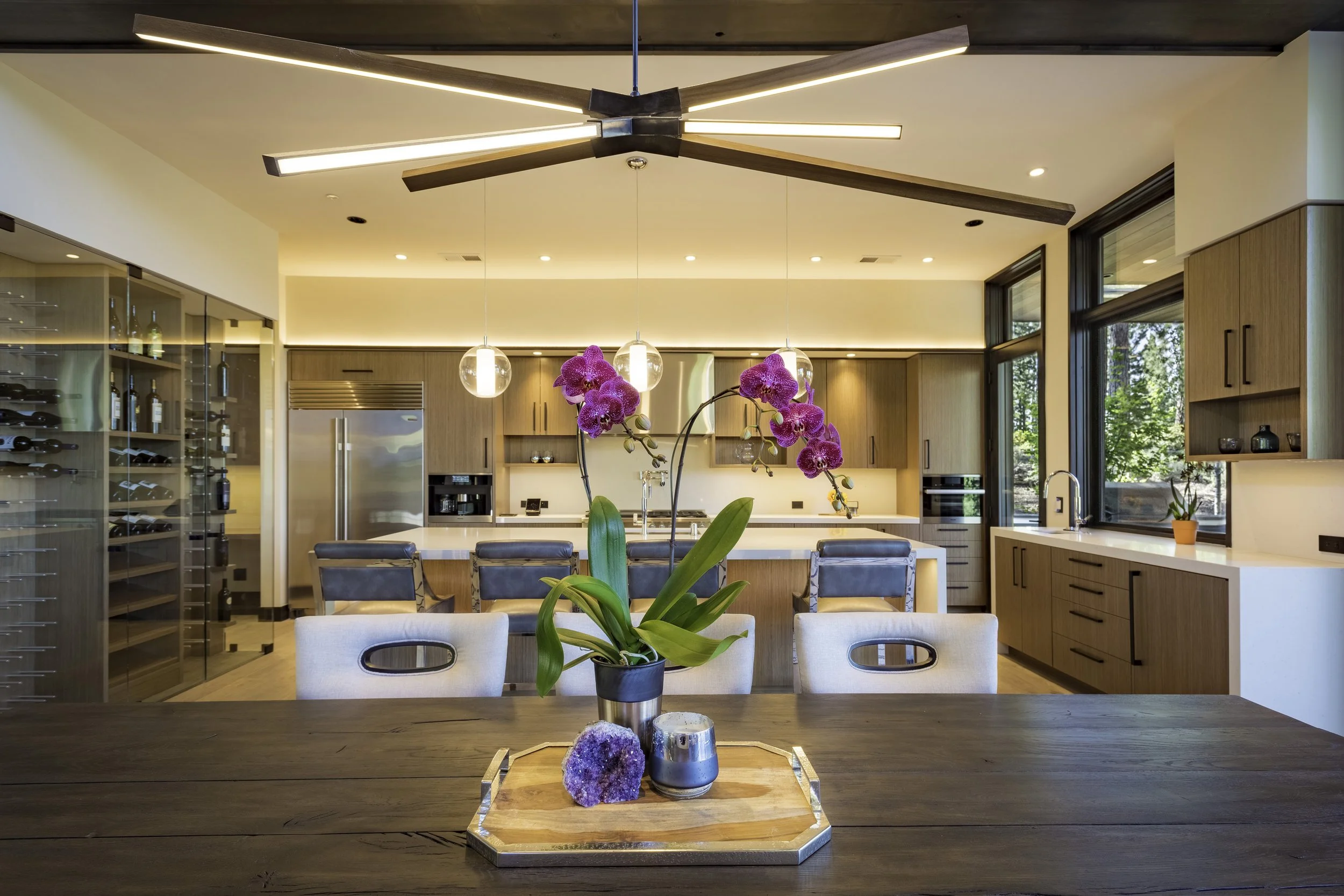 Modern kitchen with wooden cabinetry, large windows, a central island, a dining table with orchid flowers, and wine storage on the left.