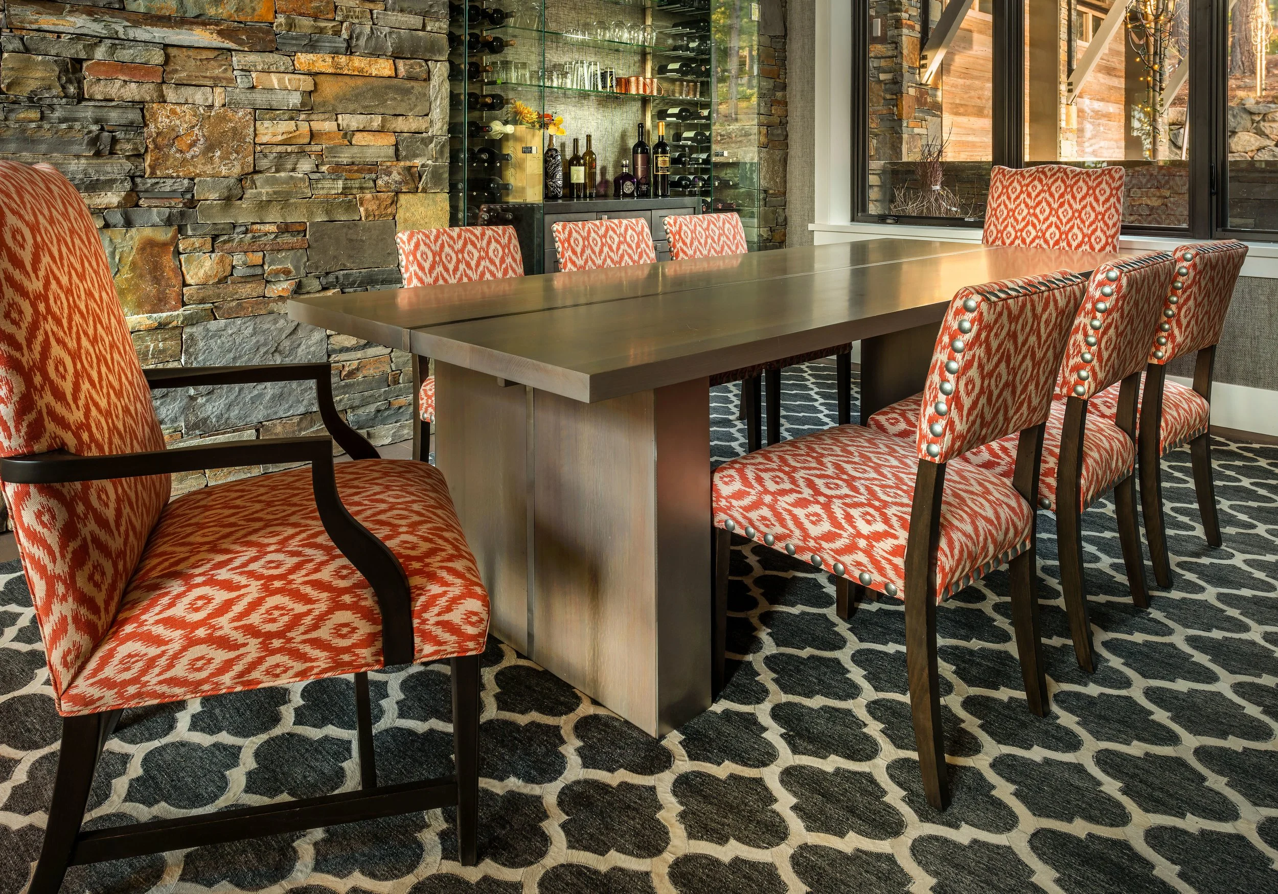 A dining room with a large wooden table surrounded by chairs upholstered in red and white geometric patterned fabric. The room has a stone accent wall, large windows, and a glass cabinet with wine bottles and glasses.