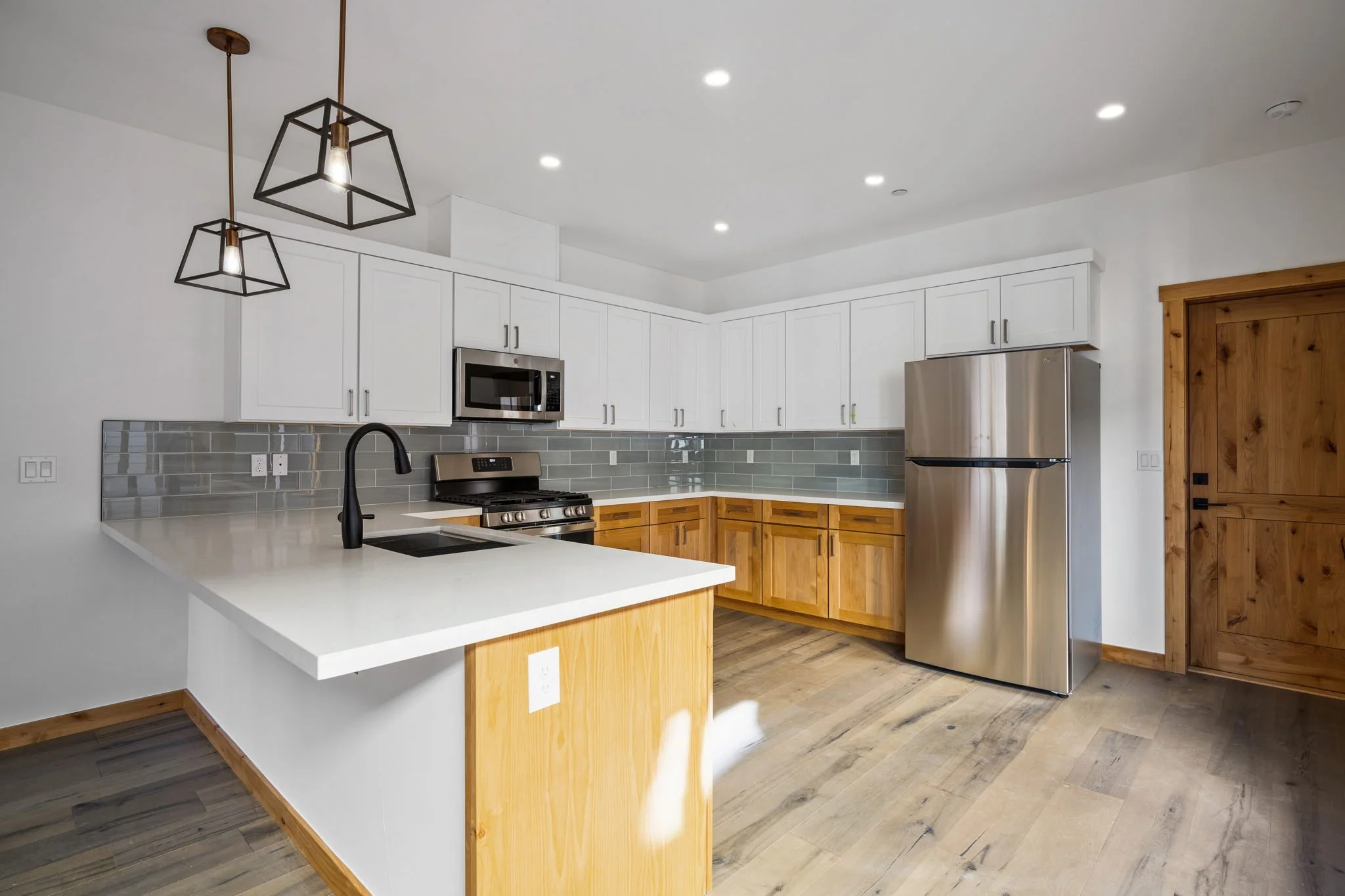 Modern kitchen with white upper cabinets, wooden lower cabinets, stainless steel refrigerator and microwave, black faucet, gray tile backsplash, light wood flooring, and black pendant lights.