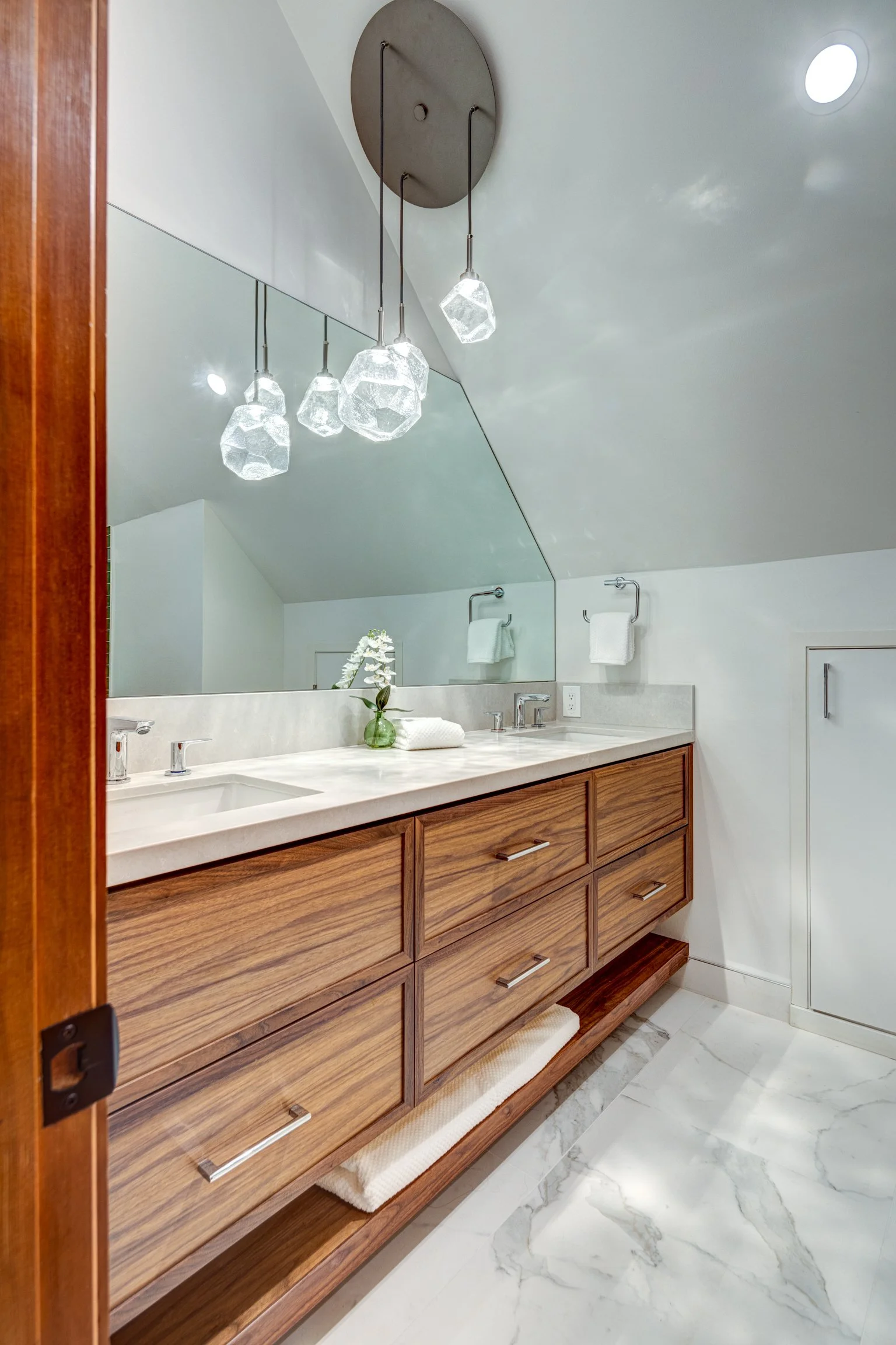 Modern bathroom vanity with wooden drawers, marble countertop, large mirror, and decorative lighting fixture. White towels and small green plant are on the counter.