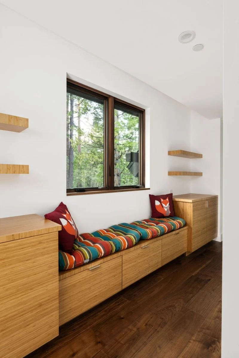 Wooden built-in window seat with colorful striped cushion, red fox-themed pillows, and wooden cabinets underneath, next to a window with a view of trees inside a modern room with white walls and dark hardwood floors.