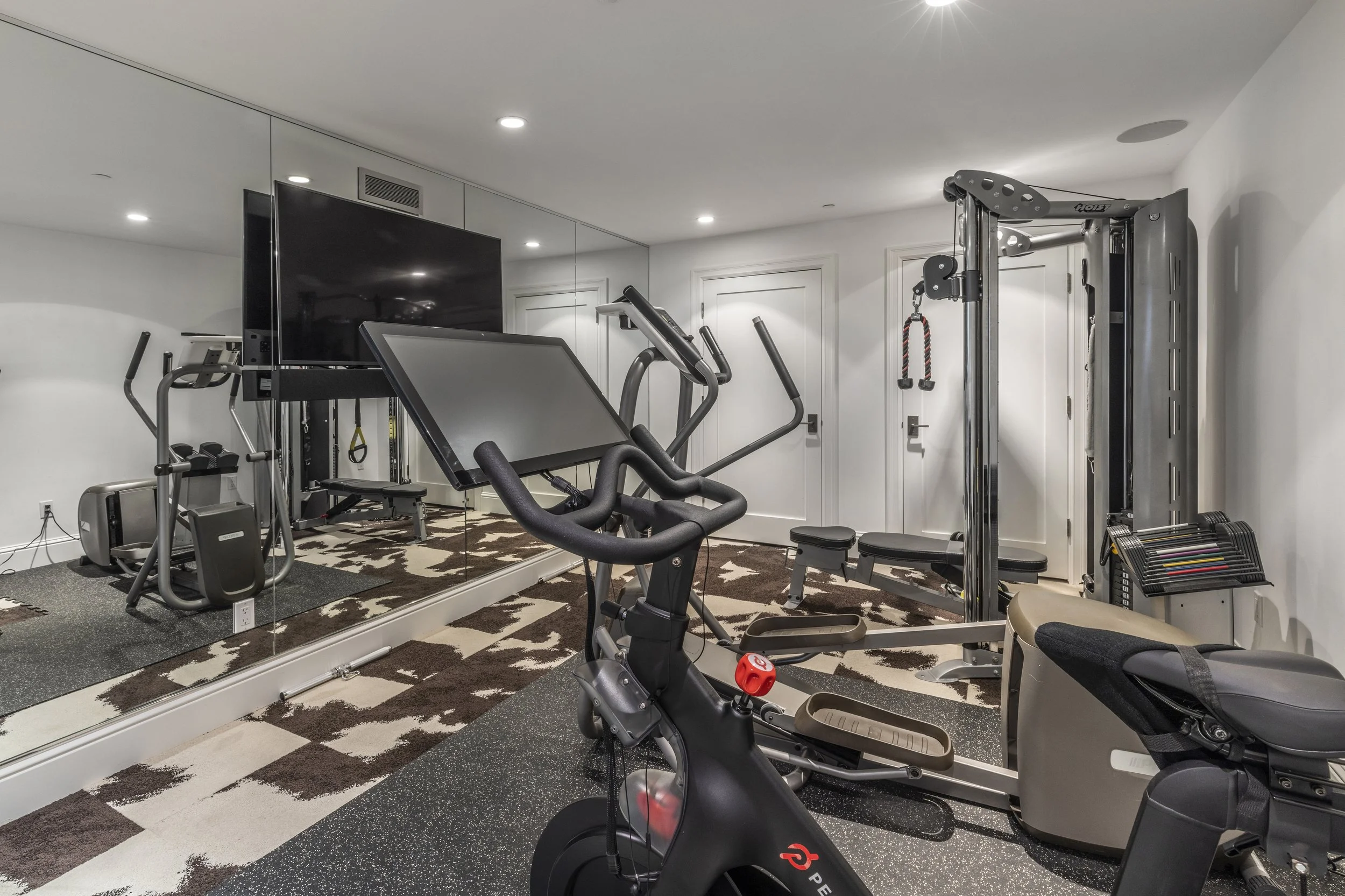 Home gym with exercise equipment, a large wall mirror, and a flat-screen TV. Includes a stationary bike, treadmill, weight machine, and various fitness accessories. Carpeted floor with geometric pattern.