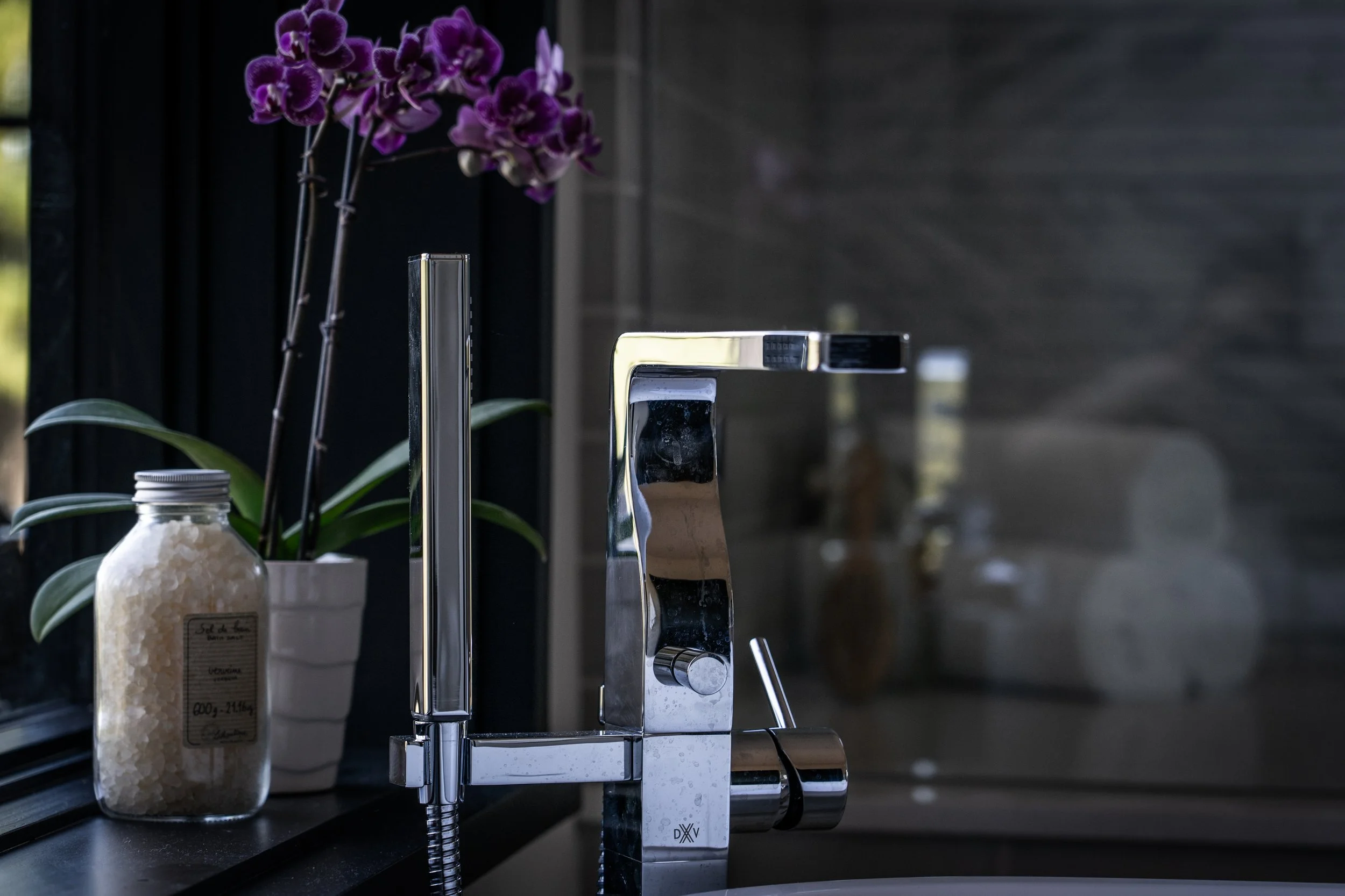 Close-up of a modern kitchen faucet, with a purple orchid plant in a white pot and a jar of bath salts on the countertop.