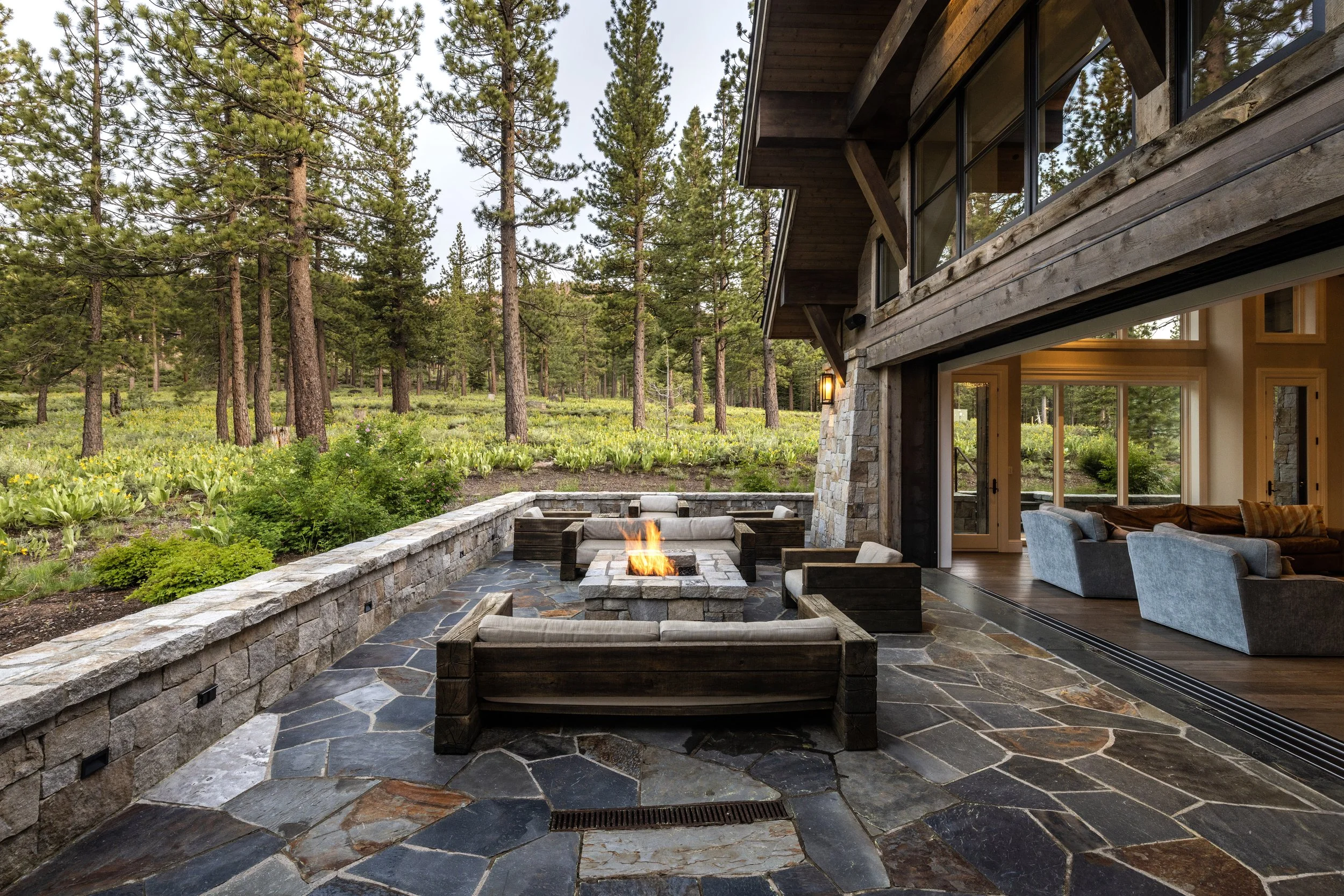 Outdoor patio with a fire pit surrounded by lounge chairs, adjacent to a house with large windows, overlooking a forested landscape with tall pine trees and greenery.